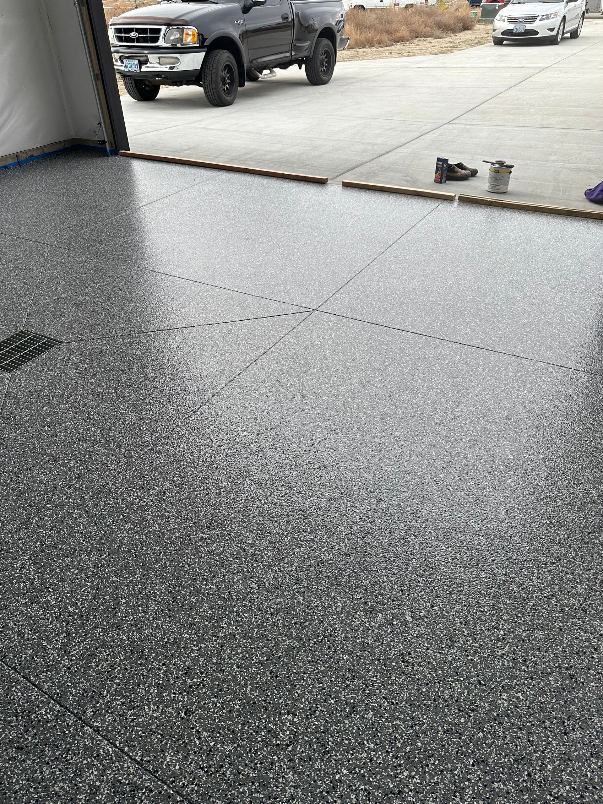 Fresh black epoxy garage floor inside open garage, with wet driveway and parked trucks outside.