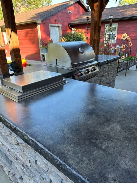 Outdoor grill station with a black countertop, barbecue grill, and red building in the background