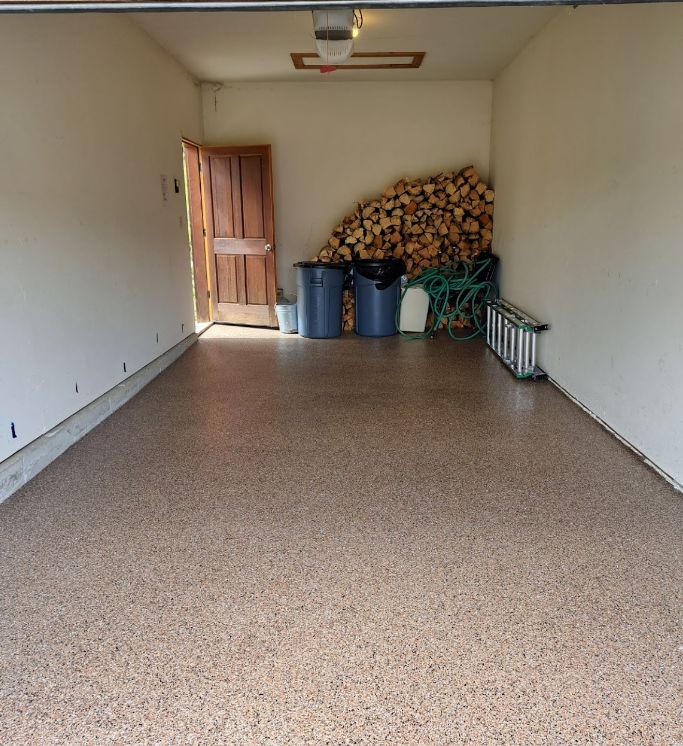 Empty garage with speckled floor, open door, and stacked firewood and bins in the back corner