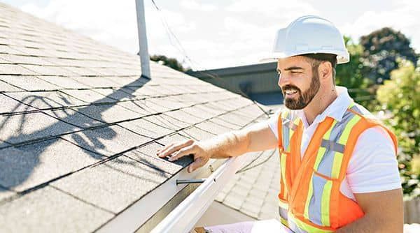 Roofer in hard hat and safety vest inspecting a roof with shingles.