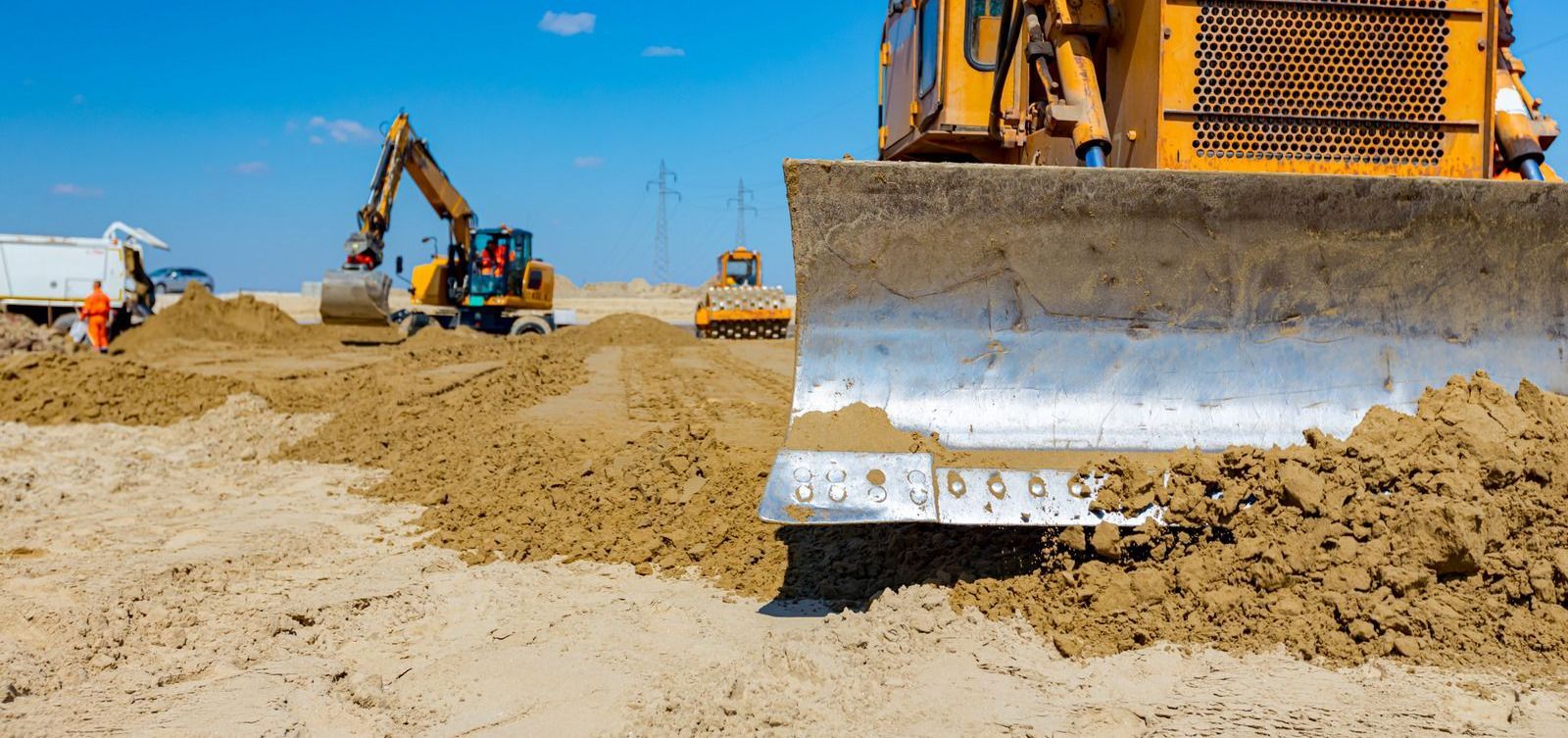 A bulldozer pushing dirt on a construction site with other machinery and workers in the background under a blue sky.