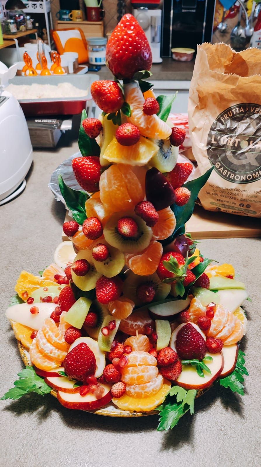 Centrotavola a forma di torre di frutta con fragole, arance, kiwi e mele, disposti su un piatto decorativo.