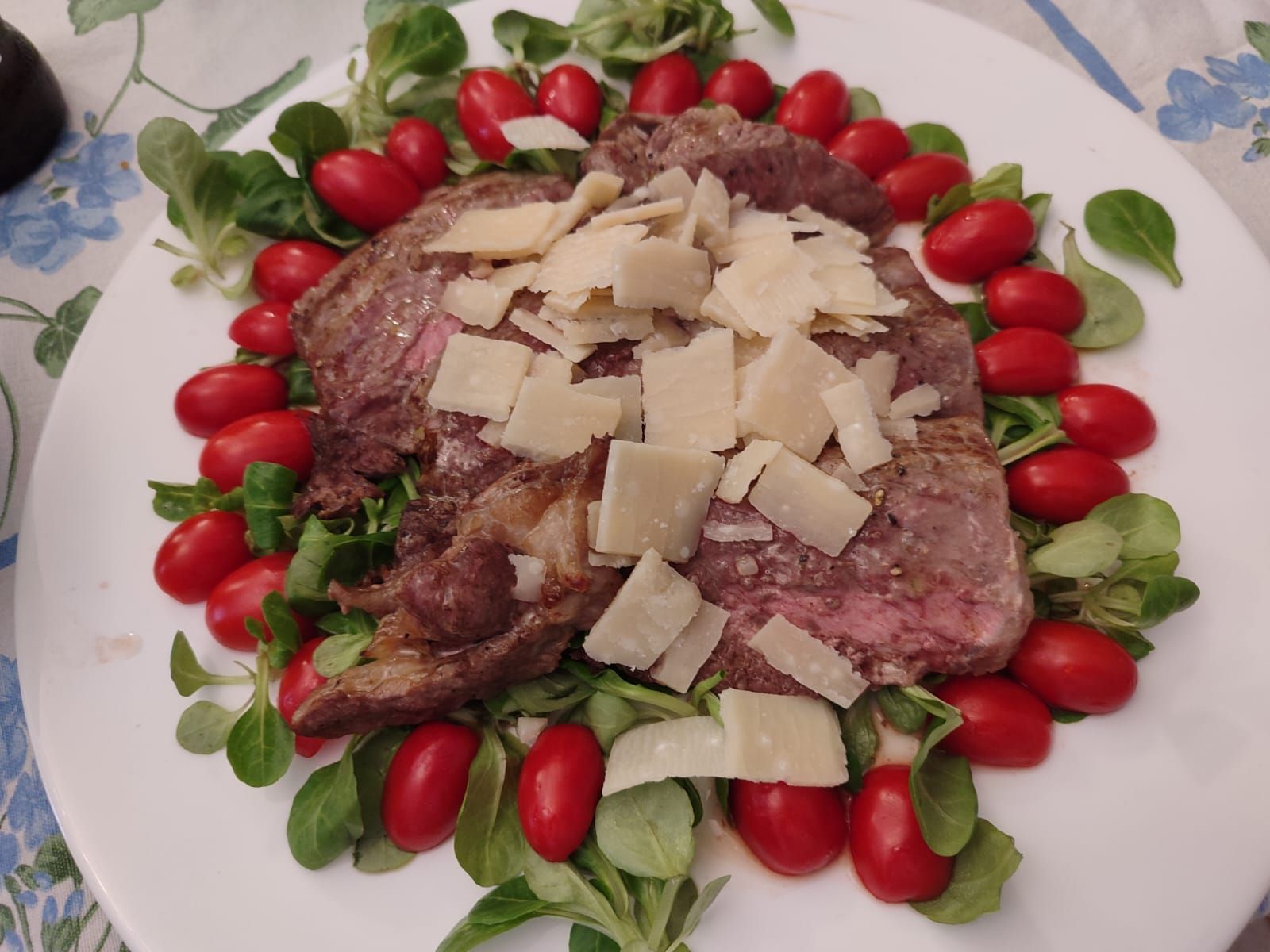Bistecca con scaglie di parmigiano, circondata da pomodori e verdure su un piatto bianco.
