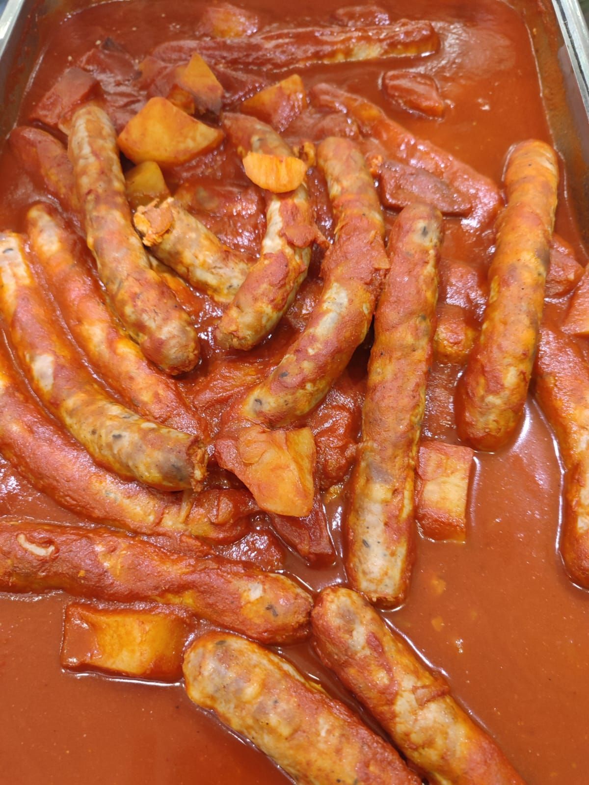 Salsicce e pezzi di patate in salsa rossa, su un vassoio da portata.