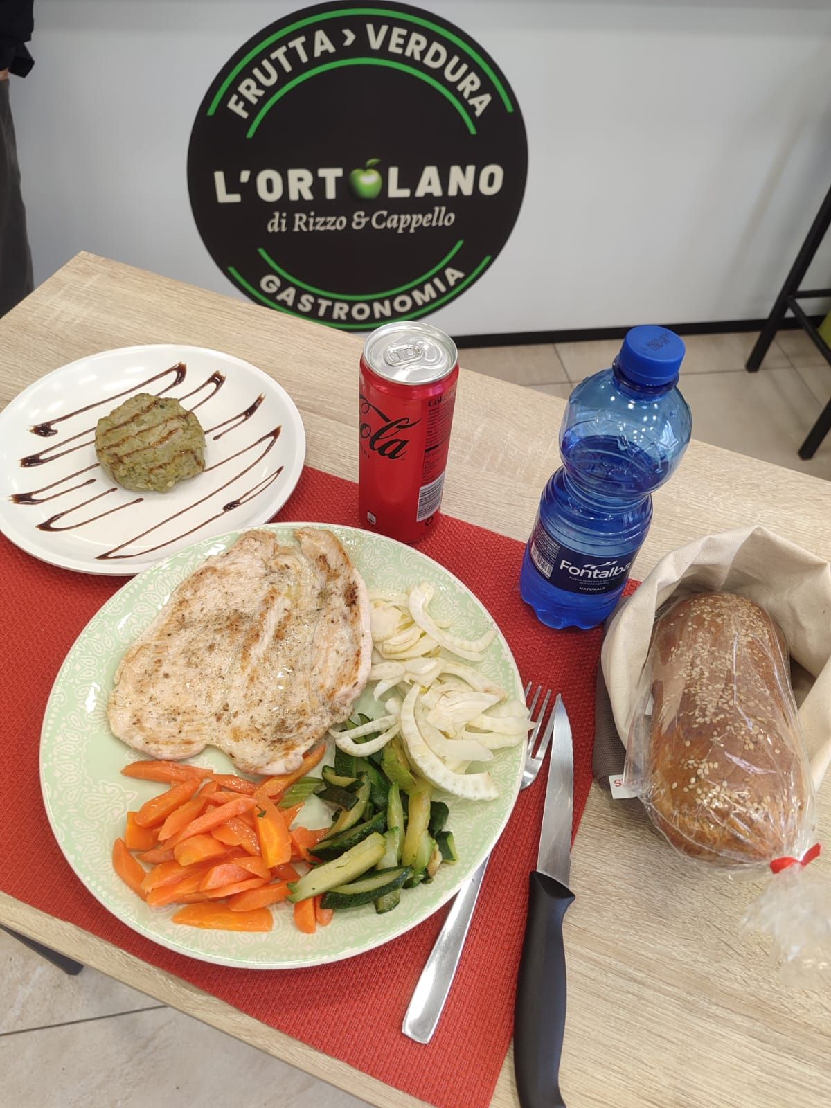 Un pasto preparato con pollo, verdure, pane, bevanda e acqua su un tavolo; logo sullo sfondo.
