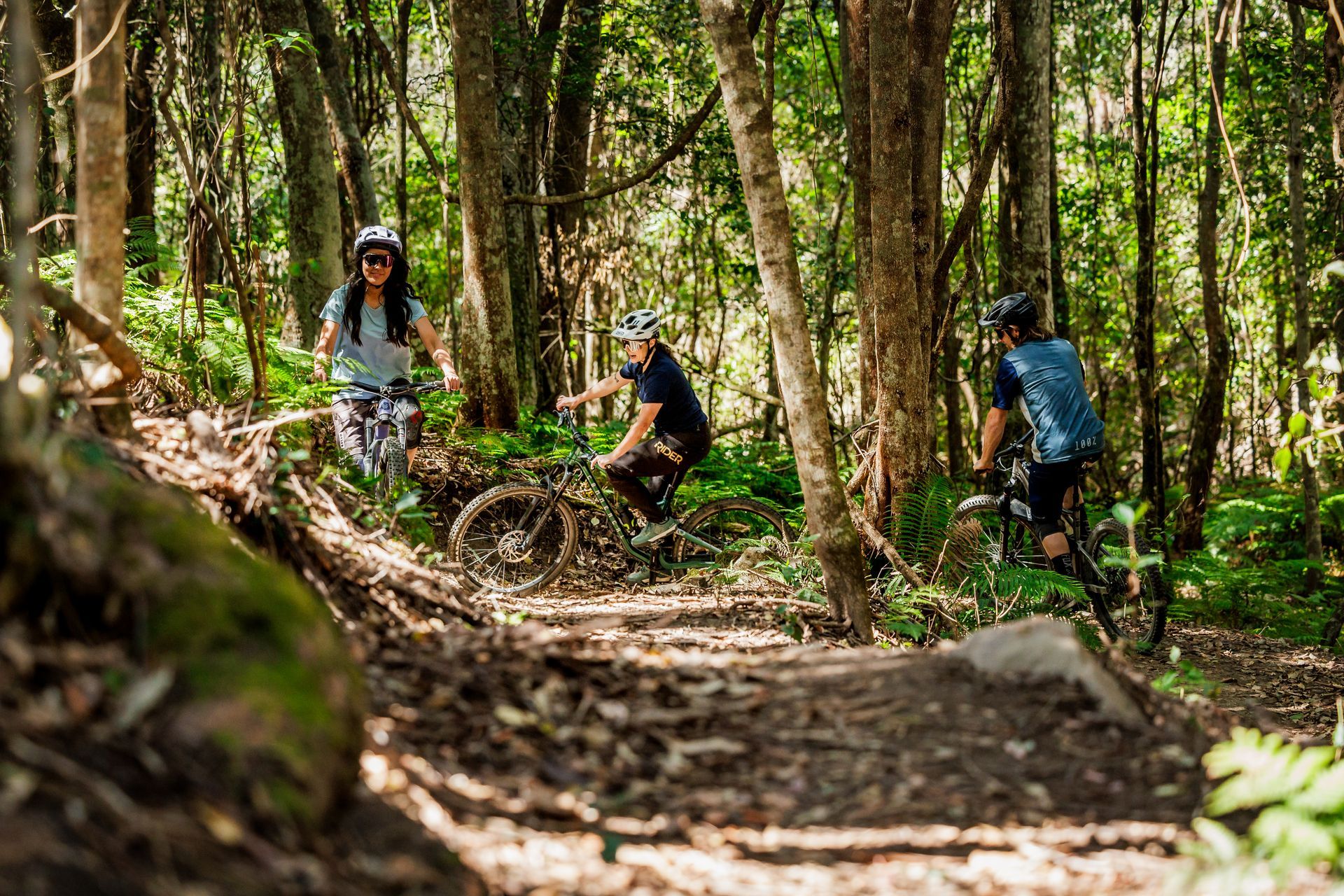 Mountain biker in red and black rides a rocky forest trail, with two riders in the background.