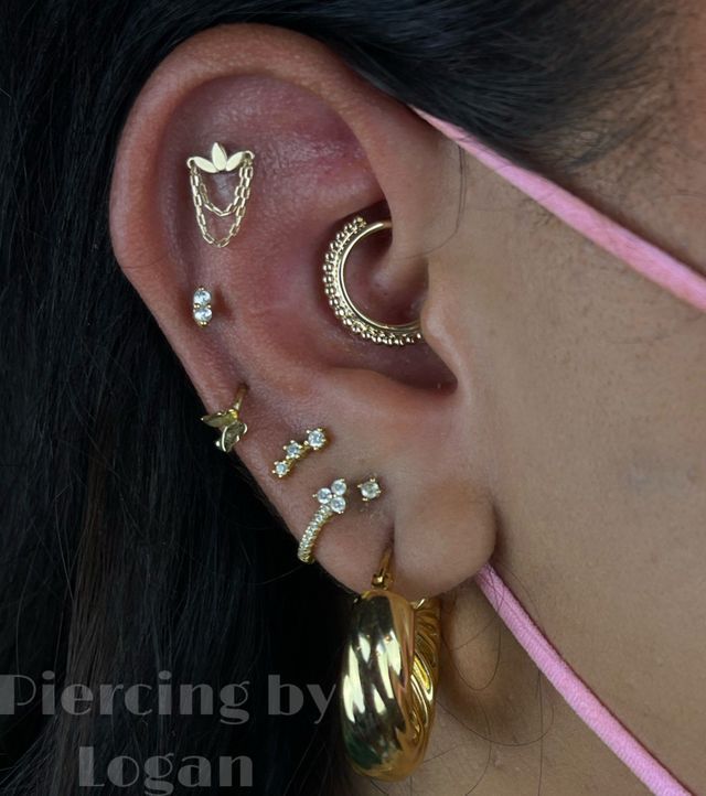 Ear with multiple gold piercings, including a crescent hoop, stud, and dangling earring.