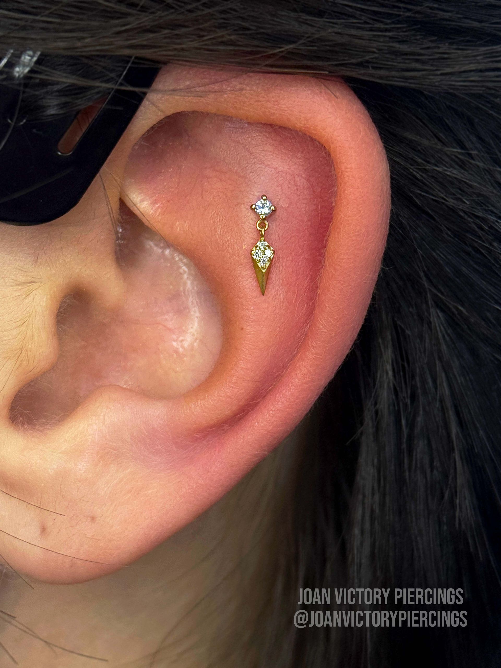 Close-up of a person's ear with a gold helix piercing. The jewelry is a gem at the top with a dangle below.