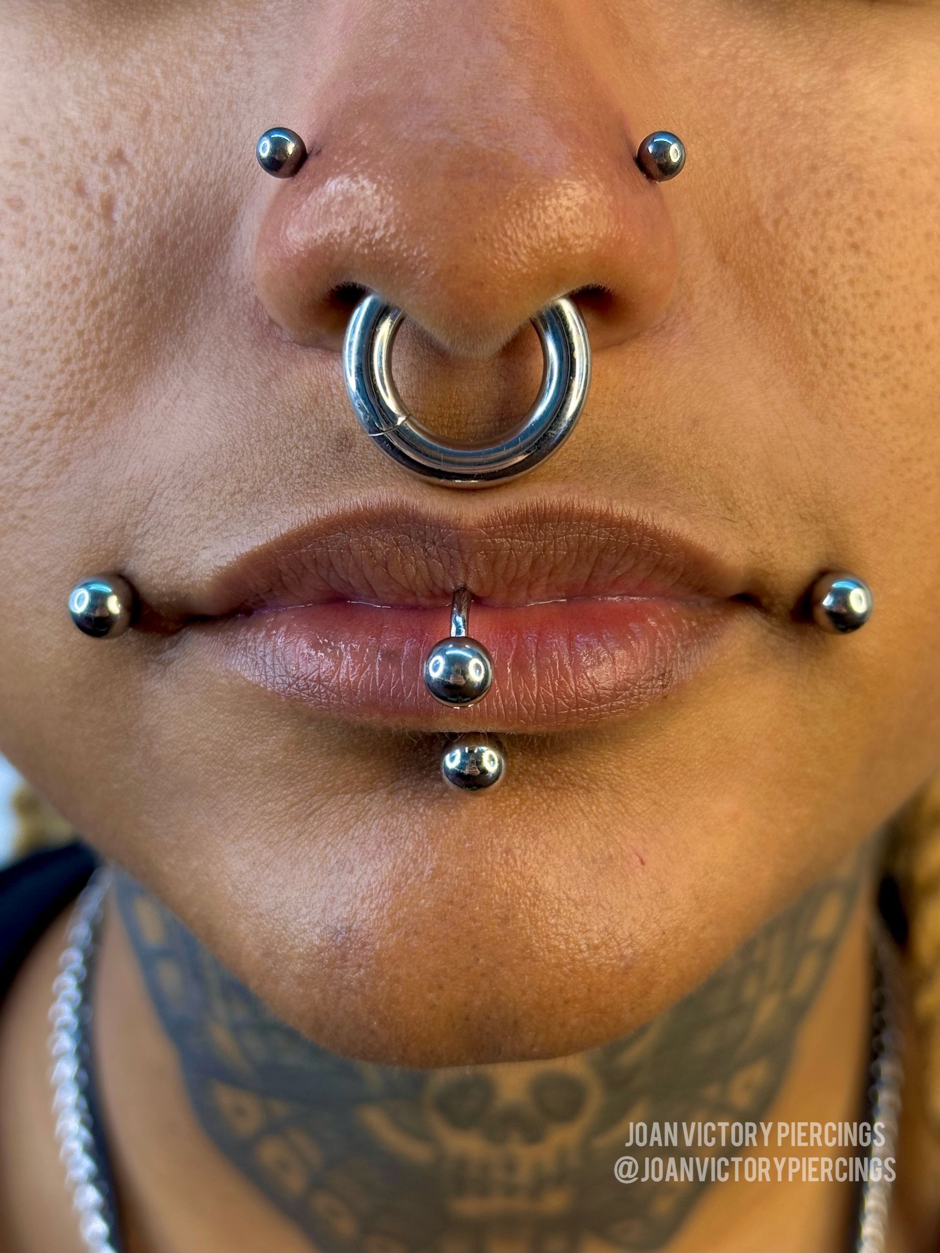 Close-up of person's face with multiple piercings: septum, nostril, lip, and snake bites. Metal jewelry.