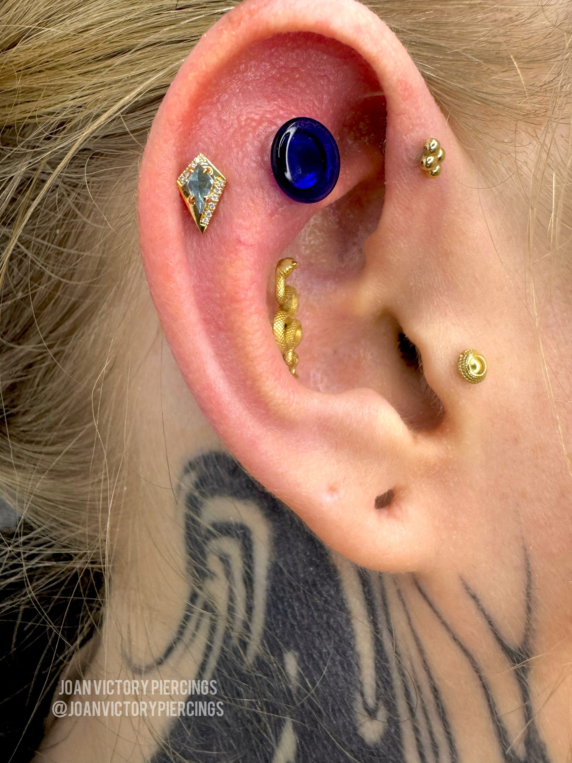 Close-up of ear with multiple piercings, including a large blue stud and gold jewelry; neck tattoo visible.