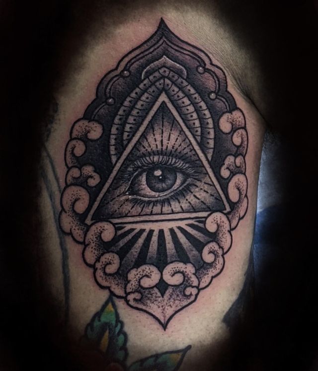 Black-and-gray tattoo of an all-seeing eye inside a triangle with ornate floral and cloud-like patterns