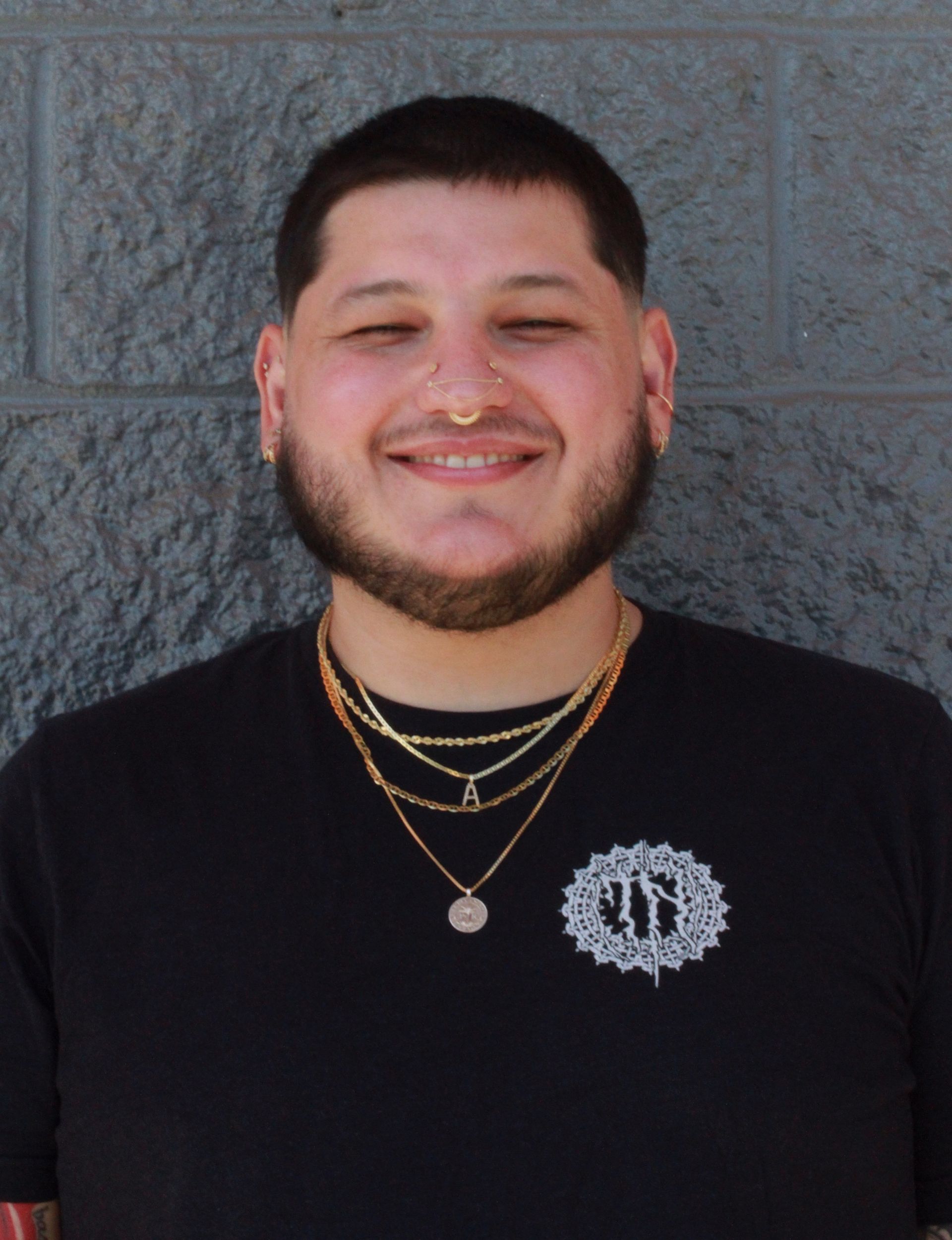 Man with short dark hair, wearing a black shirt, gold jewelry, and a nose ring, smiling.