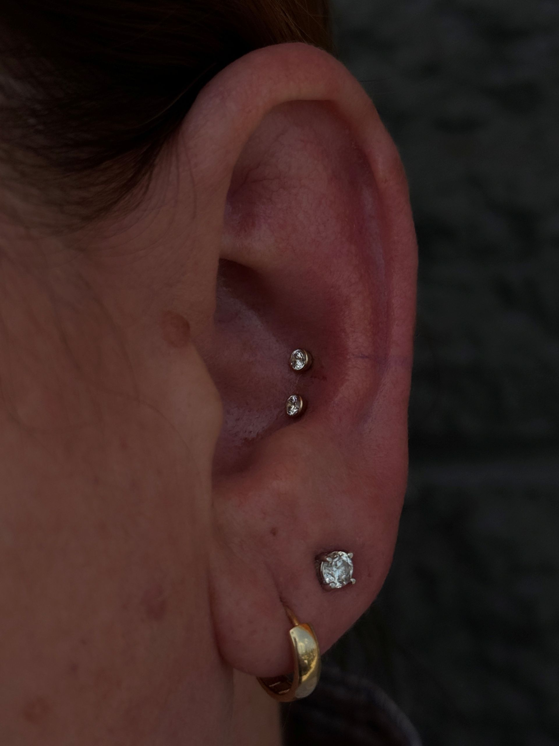 Ear with three piercings: a gold hoop, a square stud, and two small round studs in the cartilage.