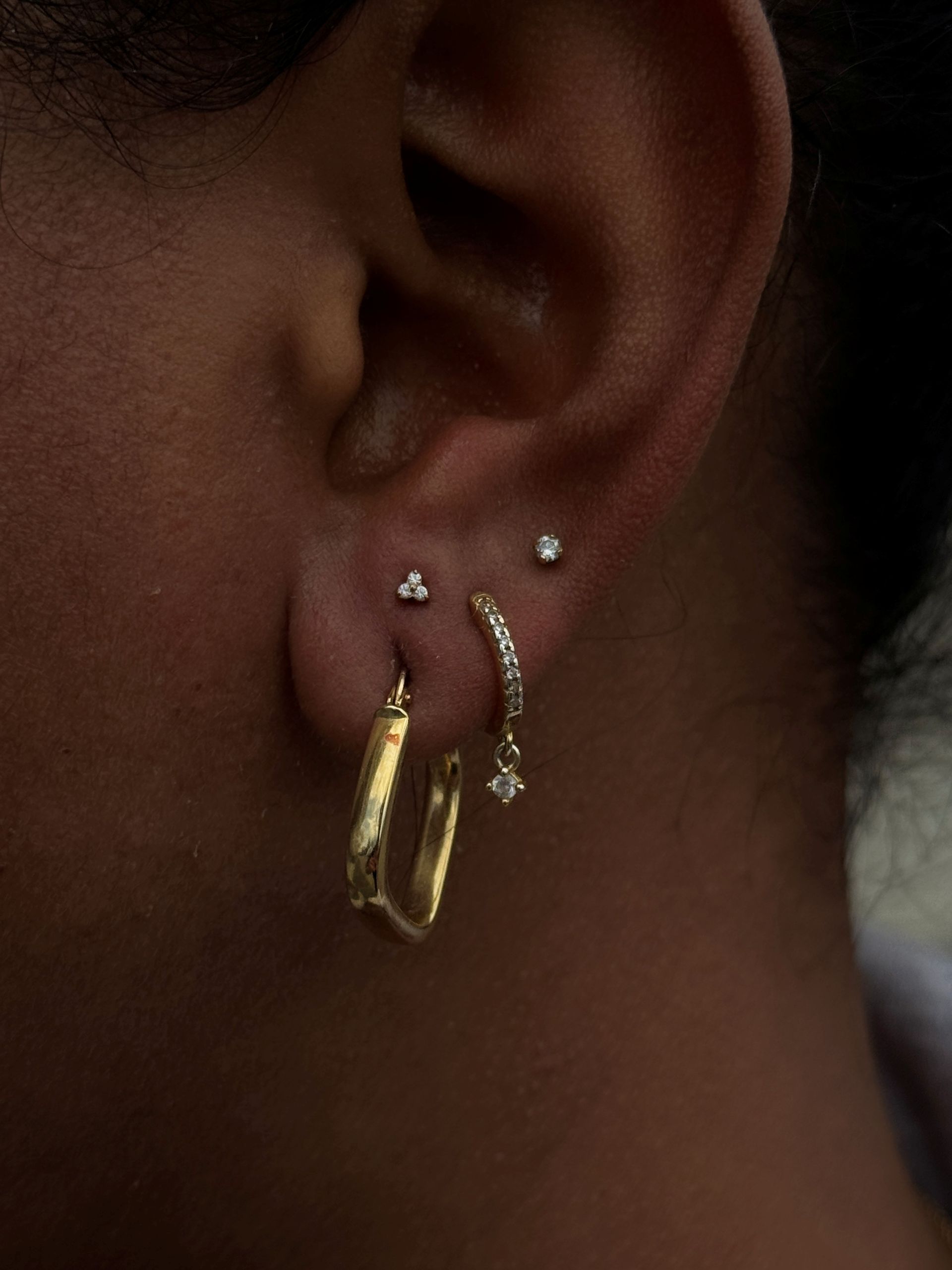 Ear with three earrings: gold geometric hoop, dangling hoop, and small stud.