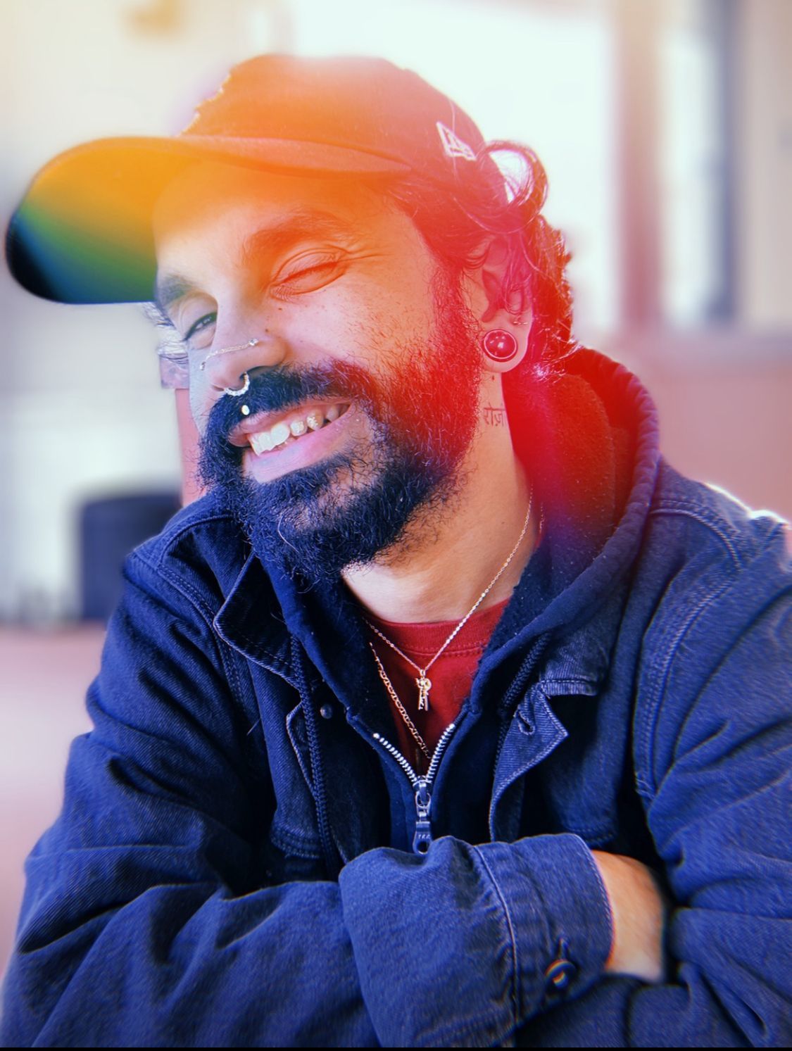 Smiling person wearing a cap, nose ring, and gauges, in a dark jacket with rainbow lens flare.