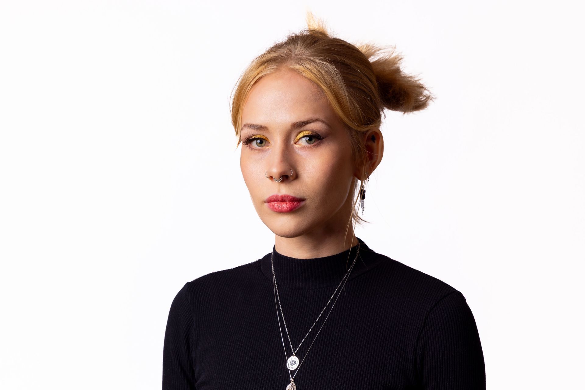 A person with light blonde hair pulled back, wearing a black high-neck top and layered silver necklaces, posing against white.