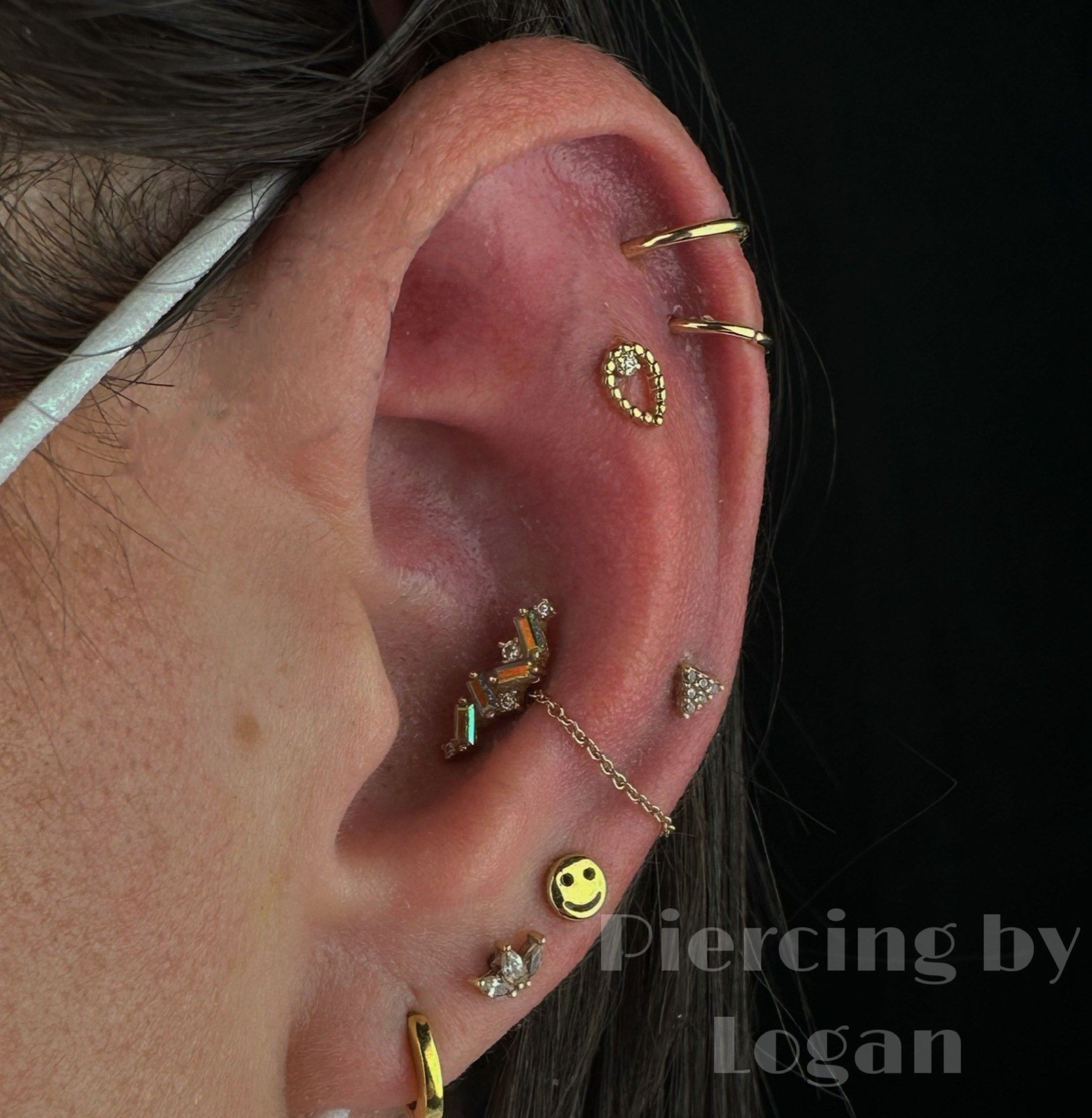 Close-up of an ear with multiple gold piercings, including a chain, and a yellow smiley face.