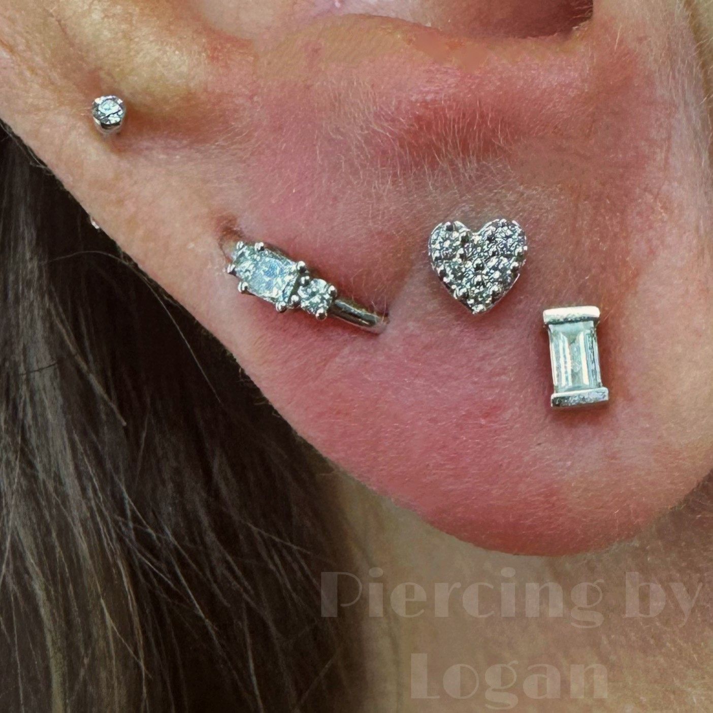 Ear with four decorative silver piercings: a stud, a bar with three stones, a heart-shaped stud, and a rectangular stud.