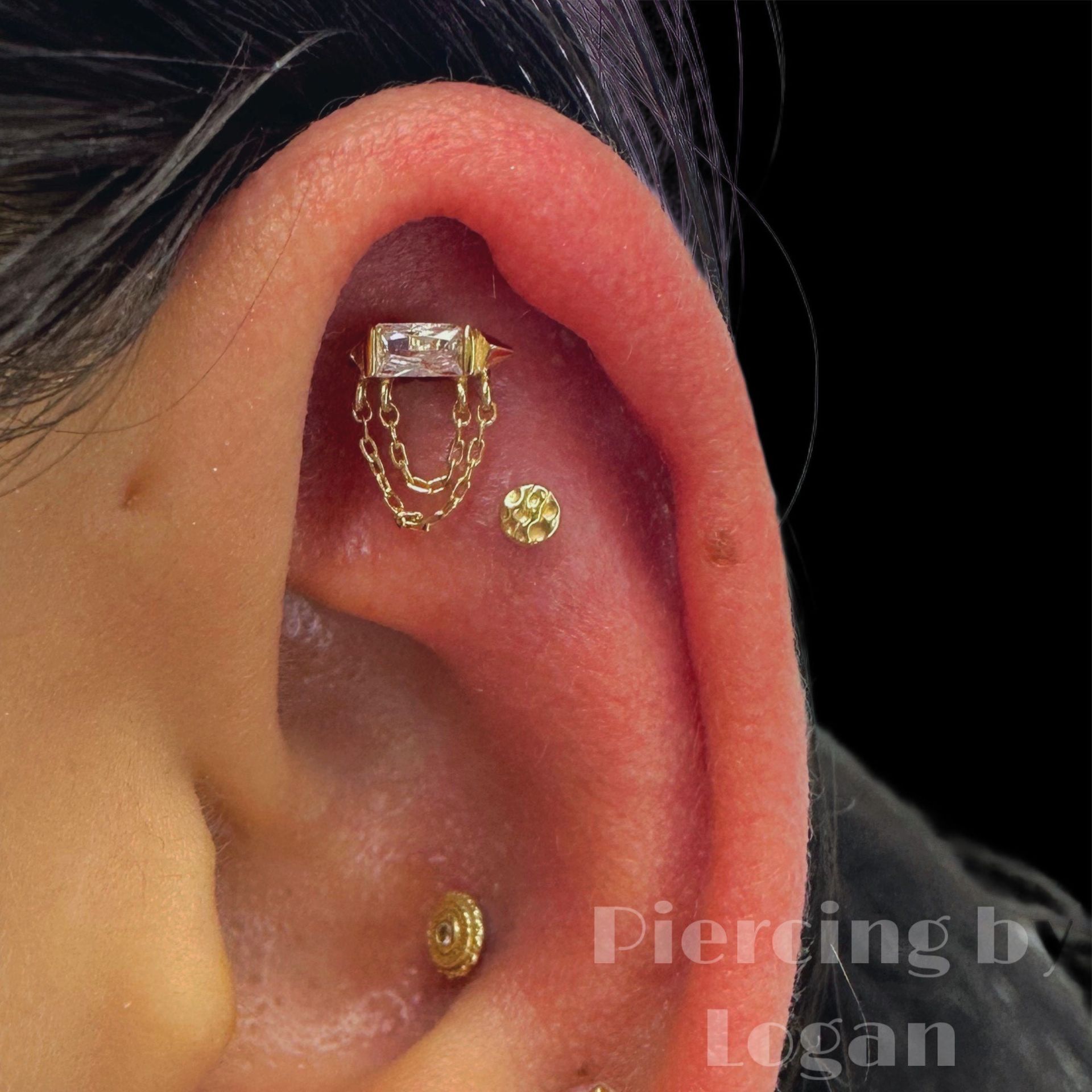 Close-up of ear with multiple gold piercings. A chain dangles from a rectangular top stud.