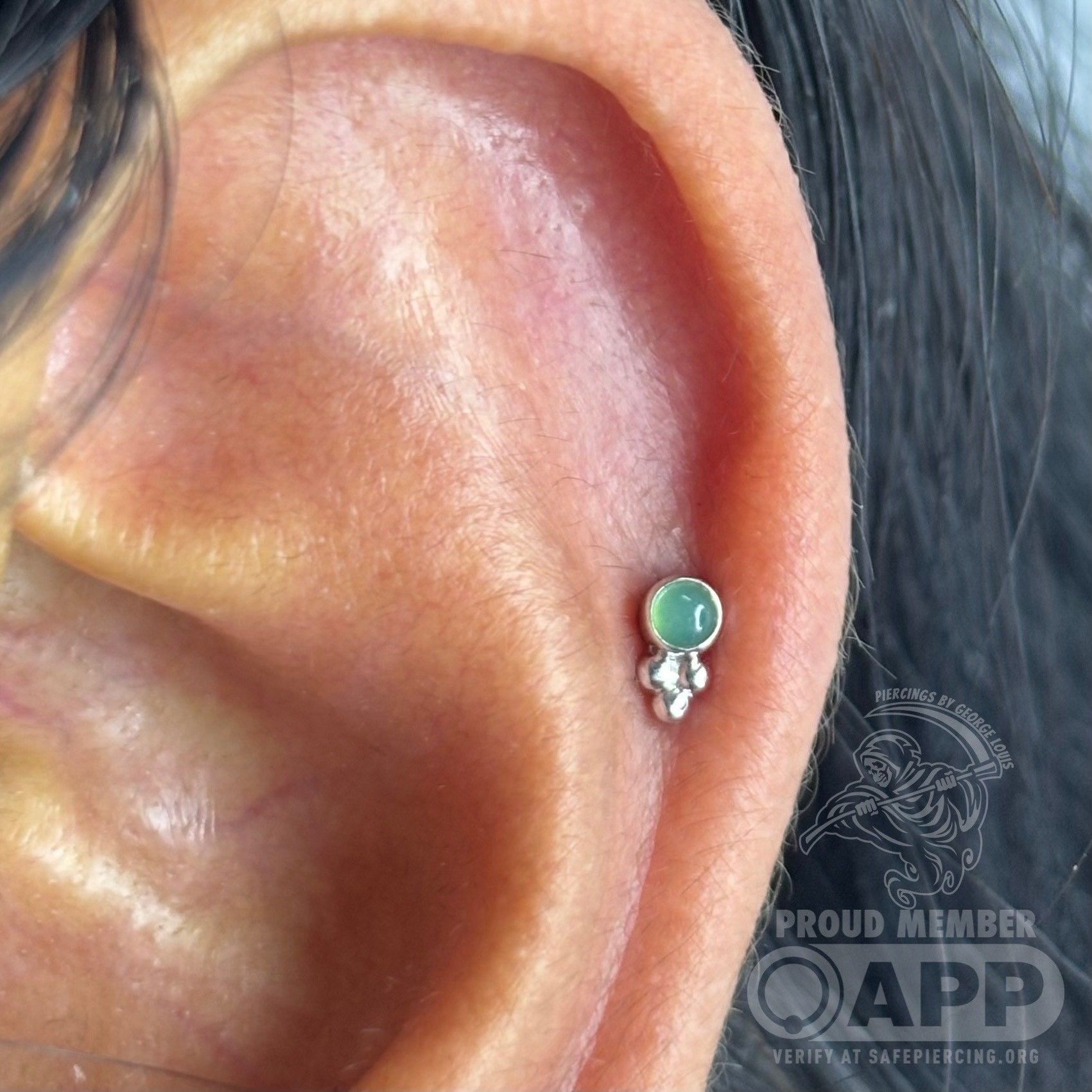 Close-up of an ear with a cartilage piercing. The piercing has a small green gem and ornate silver backing.