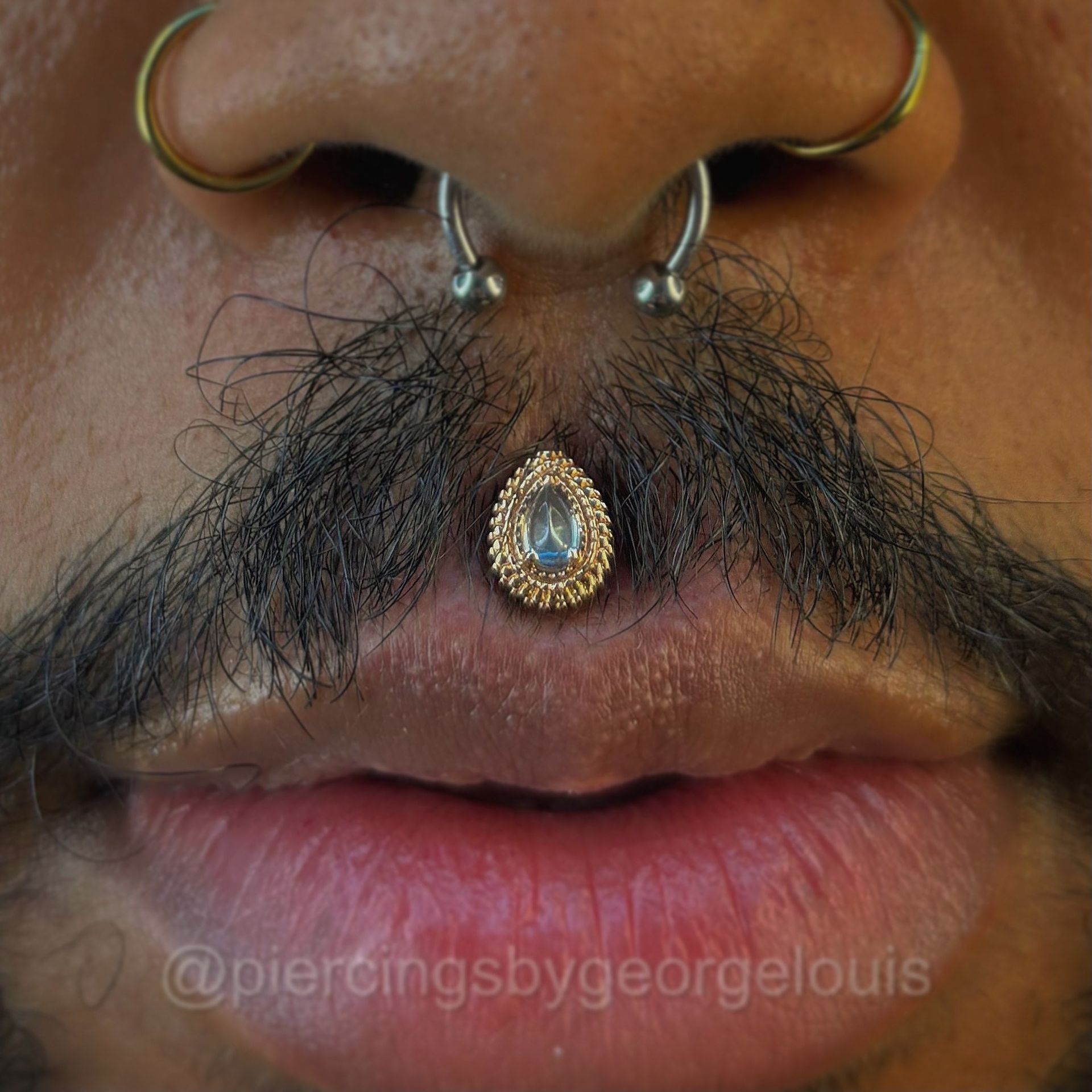 Close-up of a person's face with a teardrop-shaped philtrum piercing, gold jewelry, and a mustache. Nose piercings are also visible.