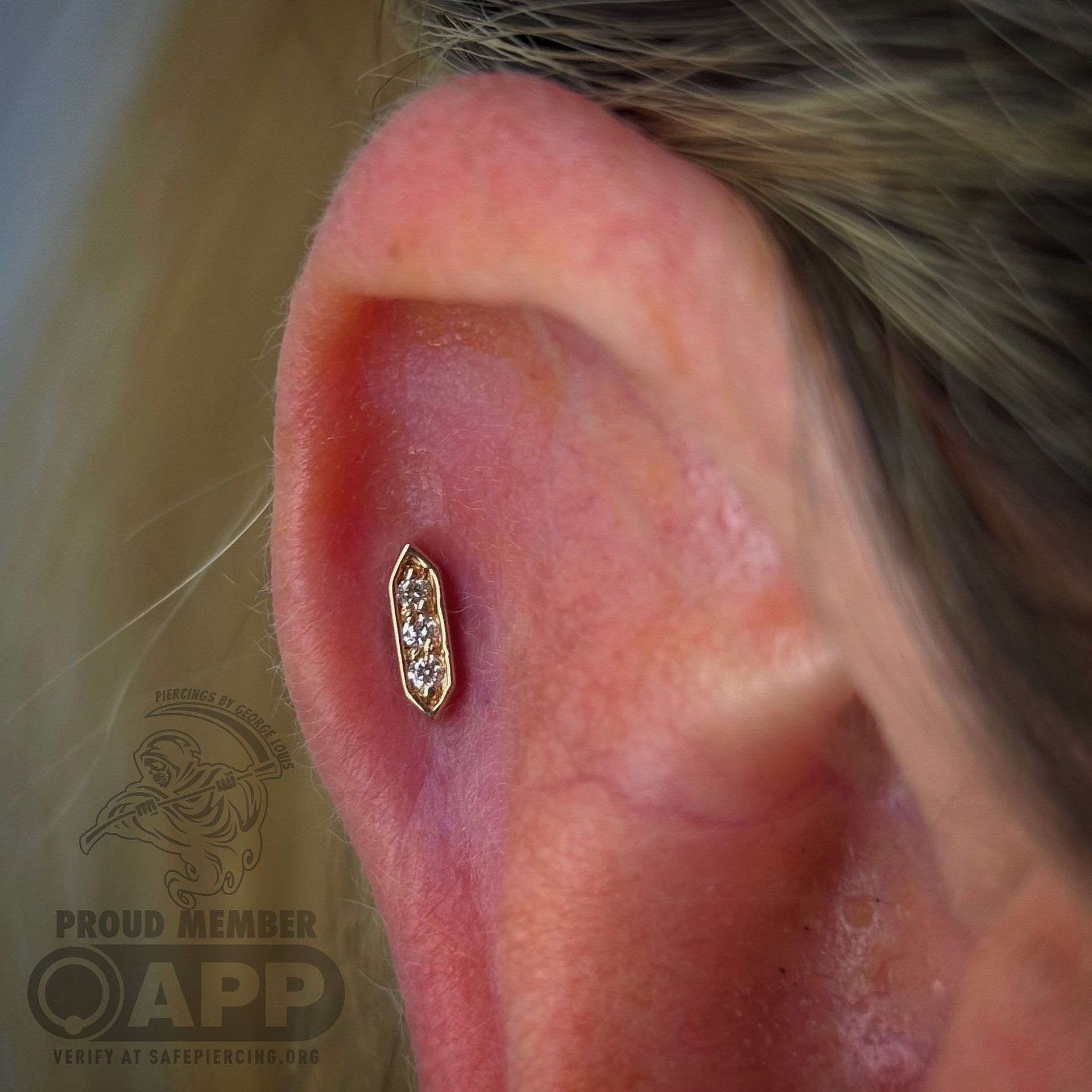 Close-up of a gold conch ear piercing with three clear gems. The ear is pink with visible veins.