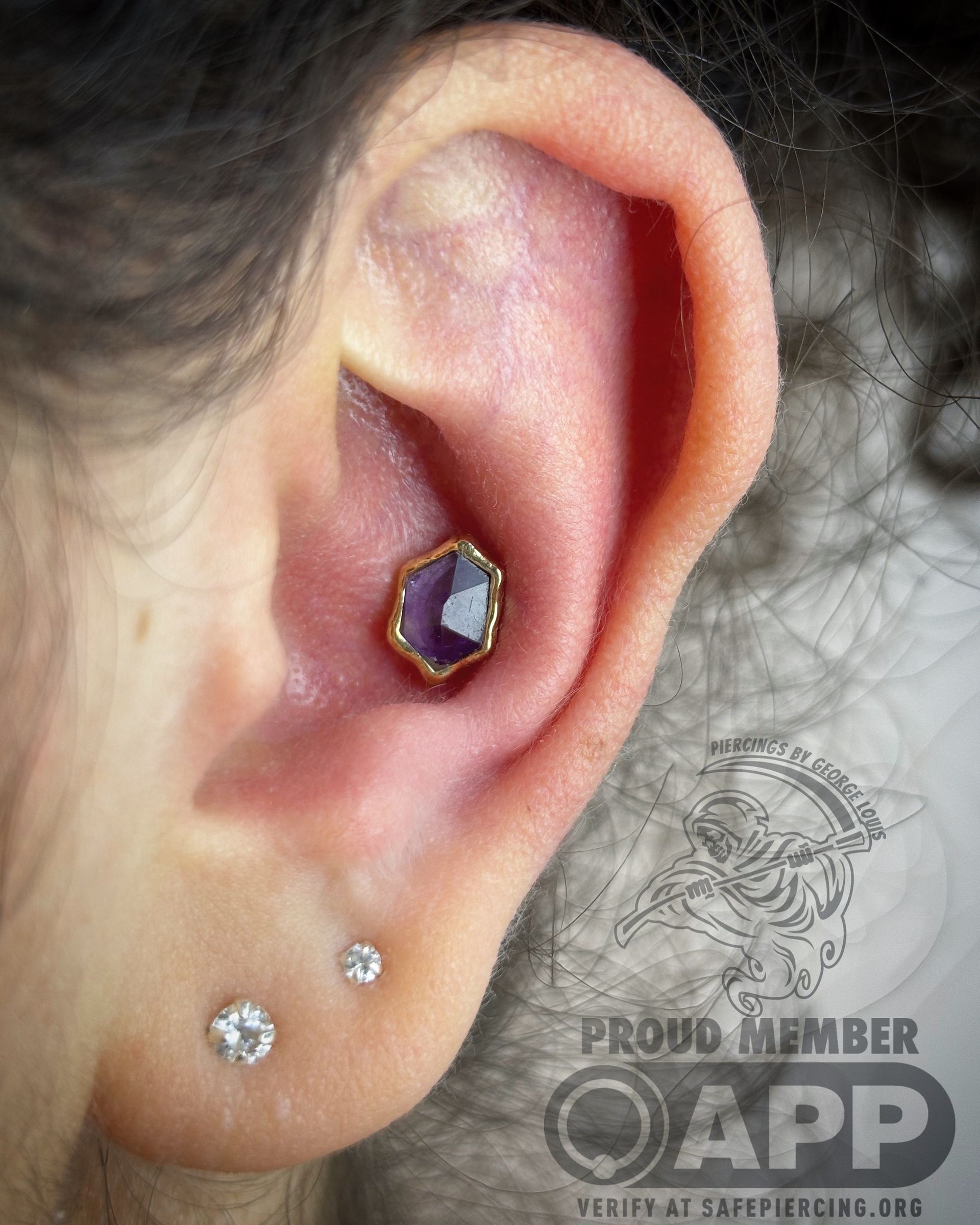 Close-up of an ear with two small piercings and a conch piercing with a hexagonal purple gem in a gold setting.