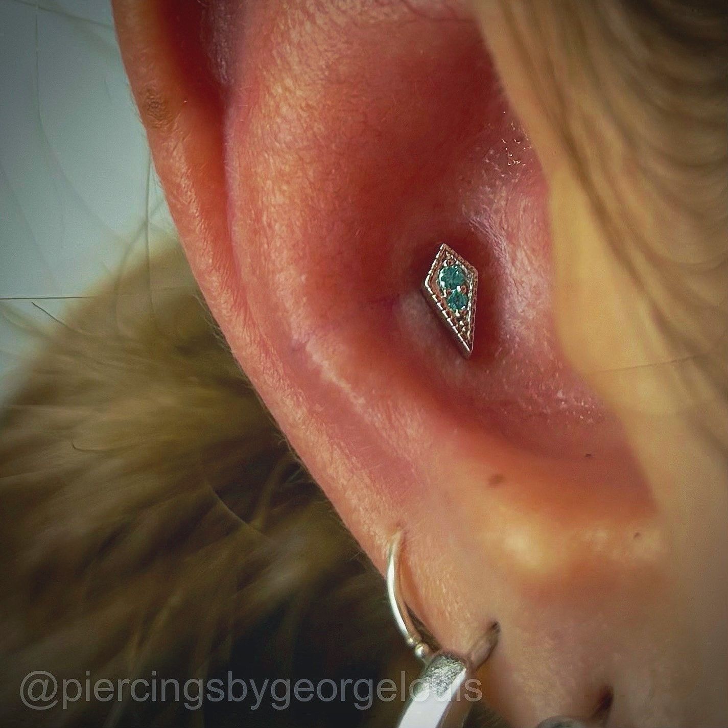 Close-up of an ear with a diamond-shaped helix piercing with teal stones; a hoop earring is also visible.
