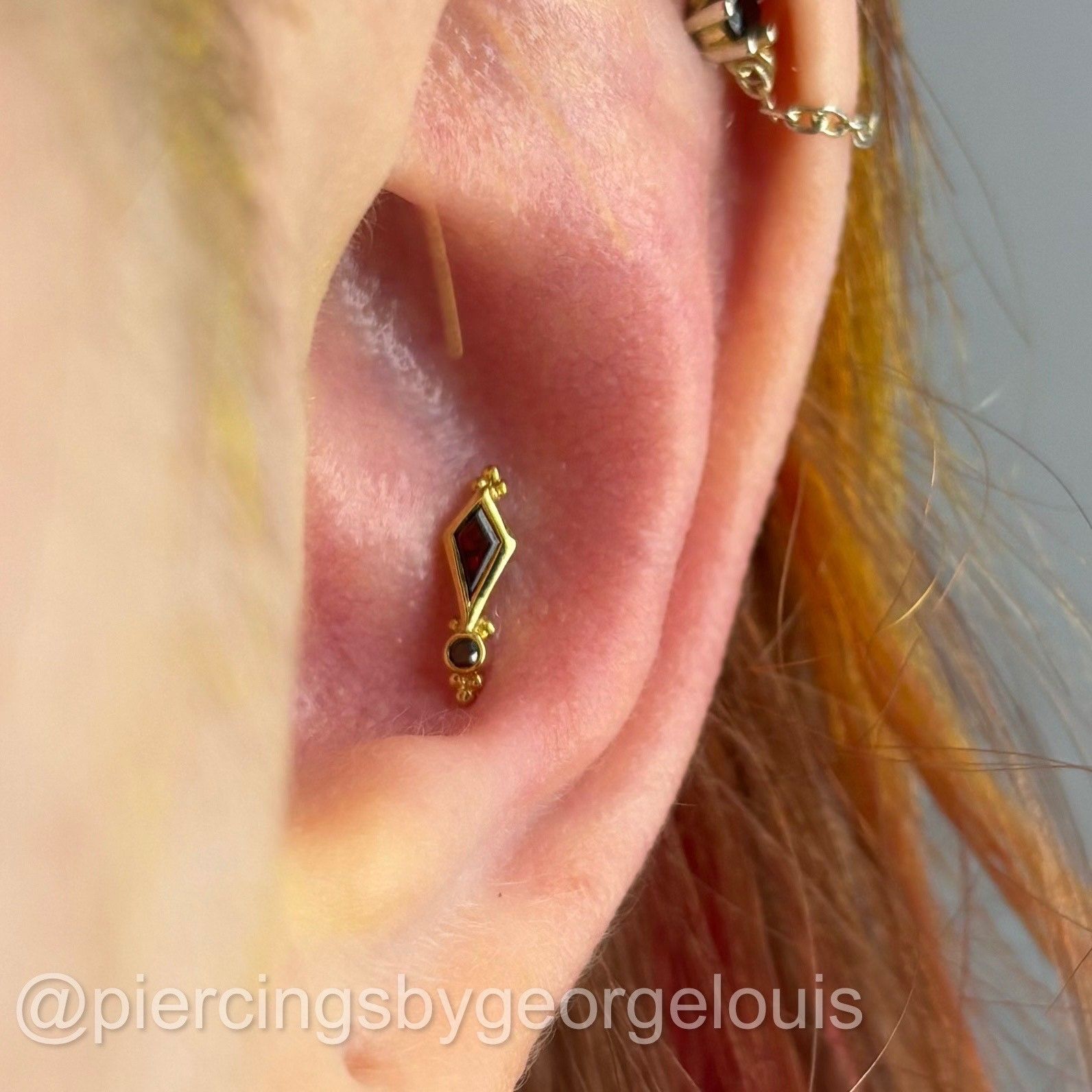 Gold and dark red ear piercing on a person's inner ear. Another piercing above is connected by a chain.
