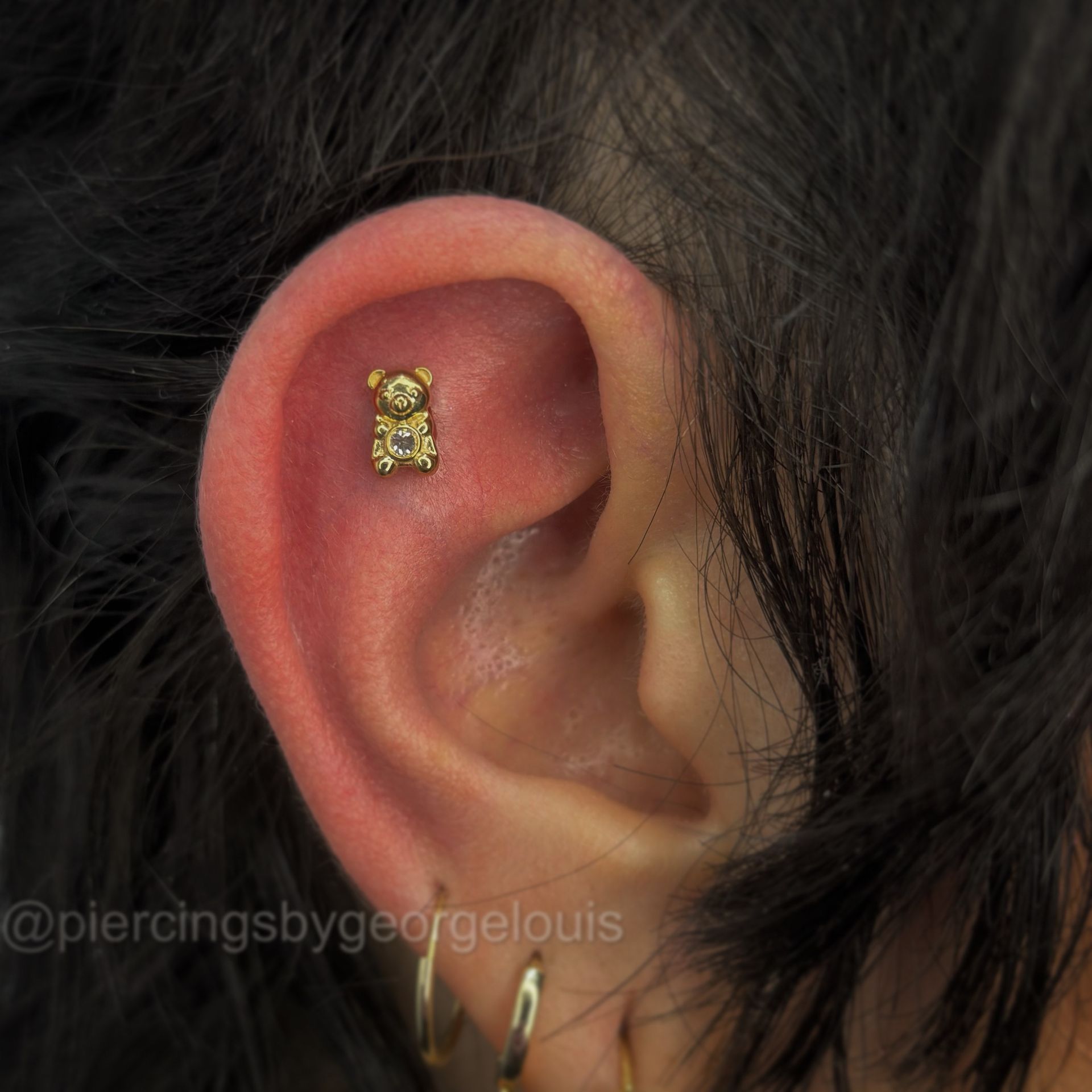 Close-up of ear with a gold teddy bear cartilage piercing, gold hoop and stud earrings.