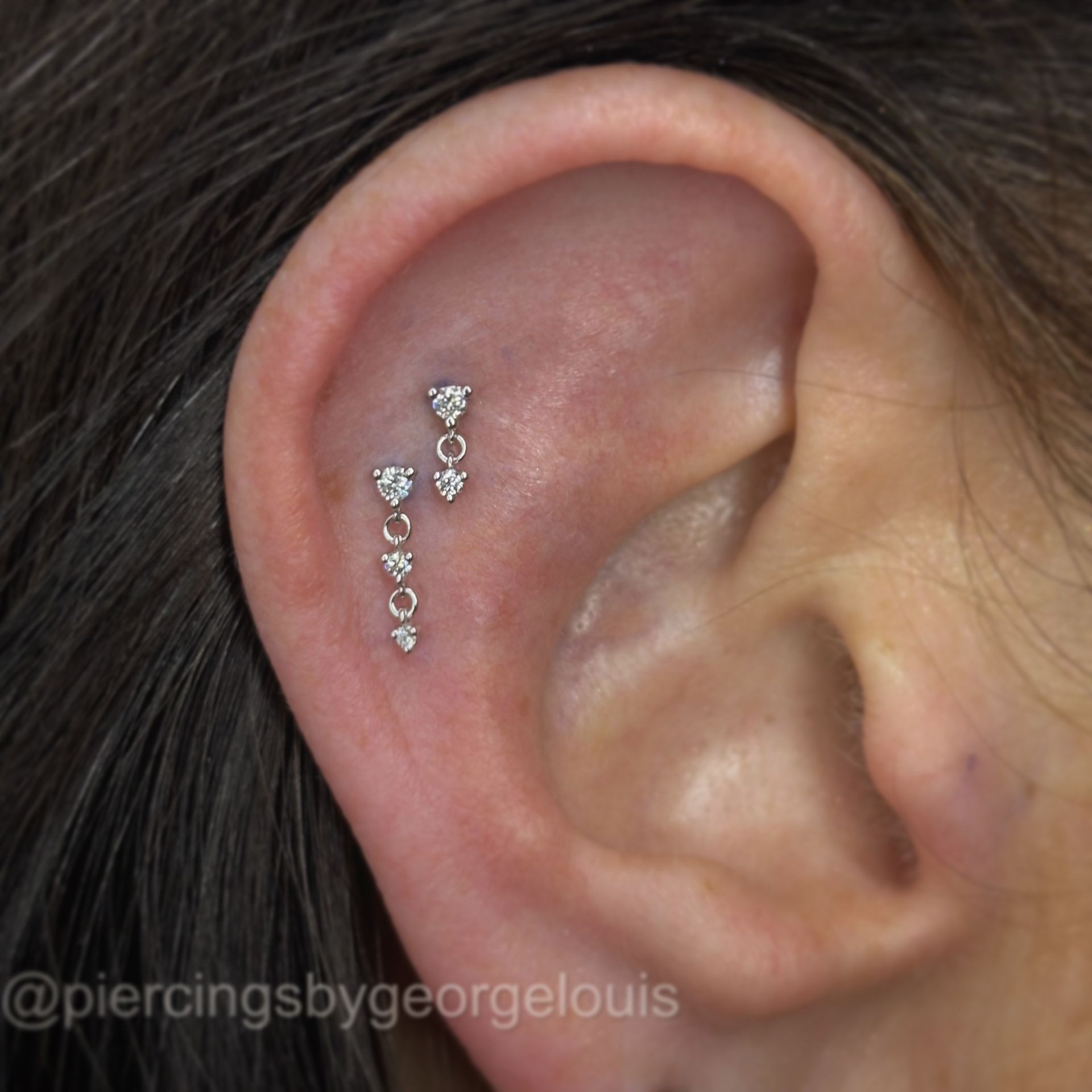 Ear with two dangling, heart-shaped jewelry piercings in the upper cartilage.