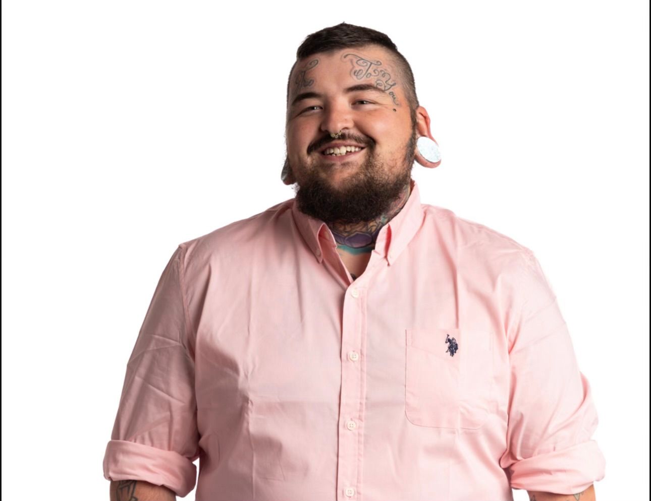 Man with tattoos and plugs smiling, wearing a pink shirt.