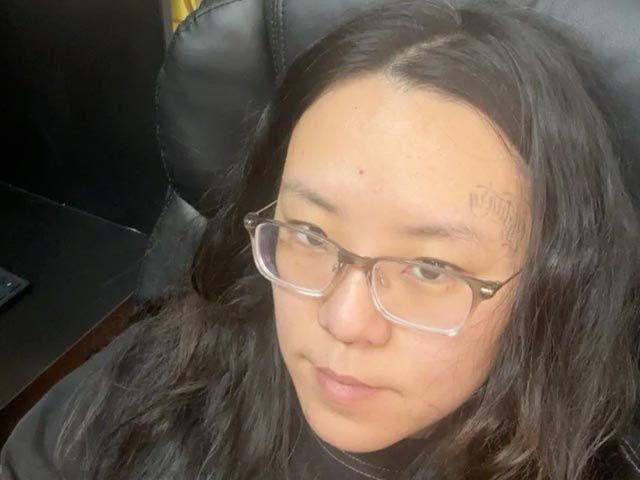 Person with glasses and long dark hair reclining on a black chair, looking at the camera.