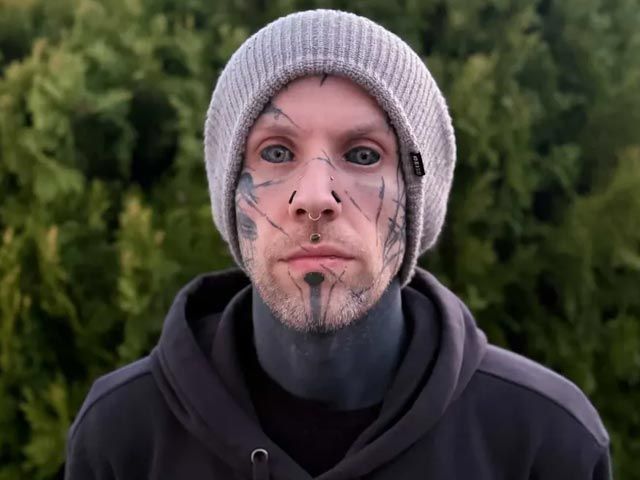 Person in a gray beanie and black hoodie, facing forward with face tattoos, against a blurred green background.
