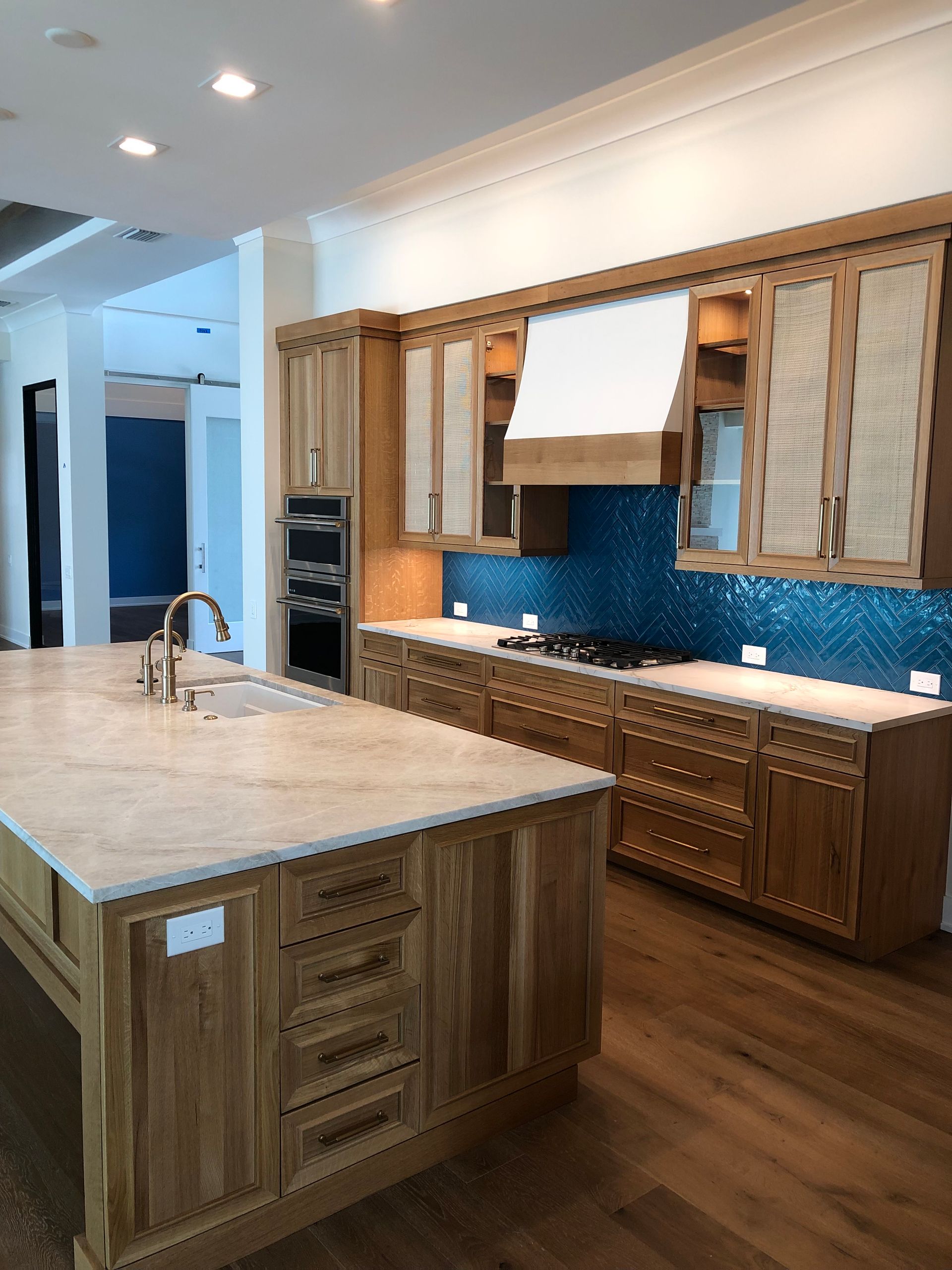 A bright kitchen with a large marble-topped island, wooden cabinetry, and a striking blue tiled backsplash.
