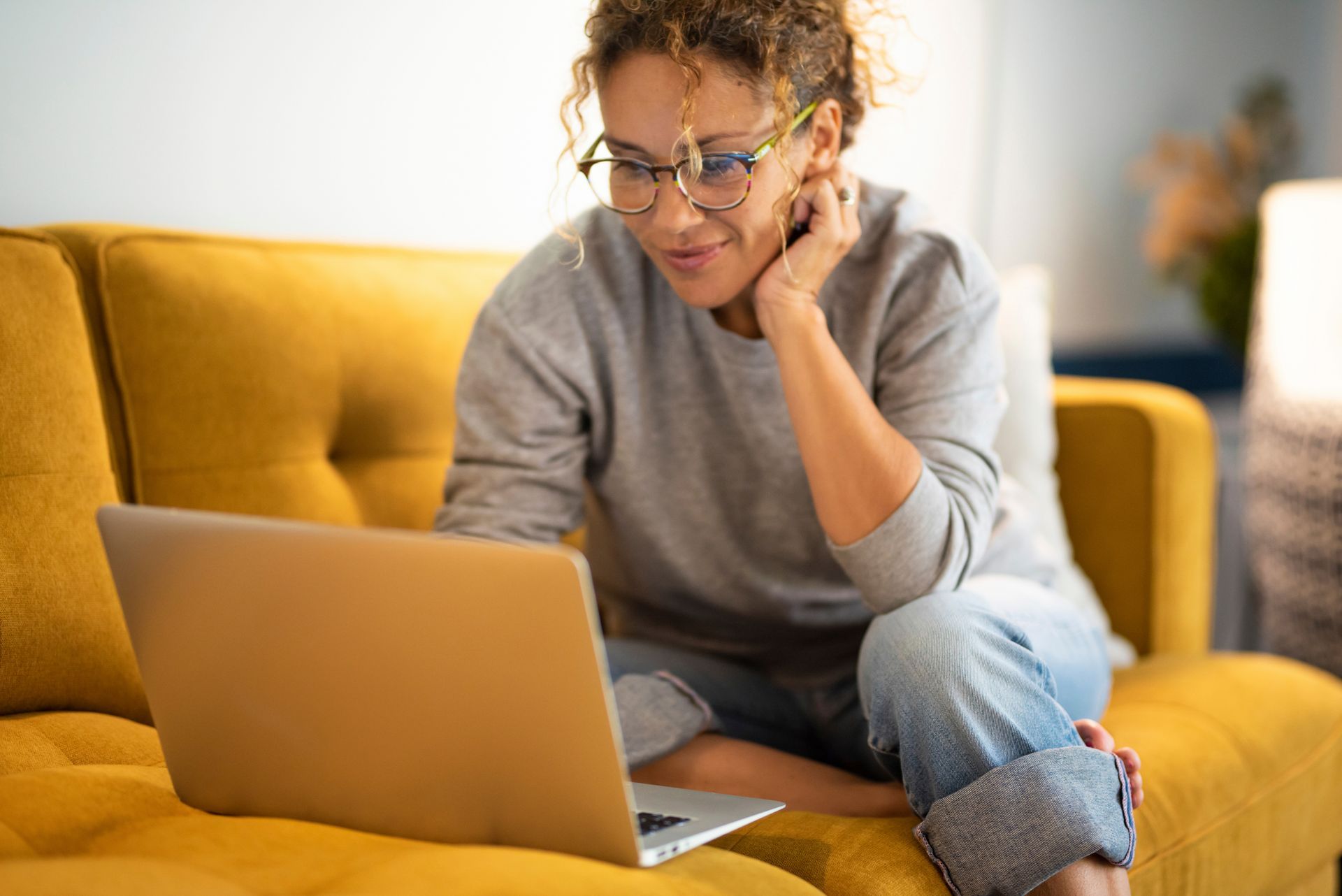 A woman is sitting on a yellow couch using a laptop computer.
