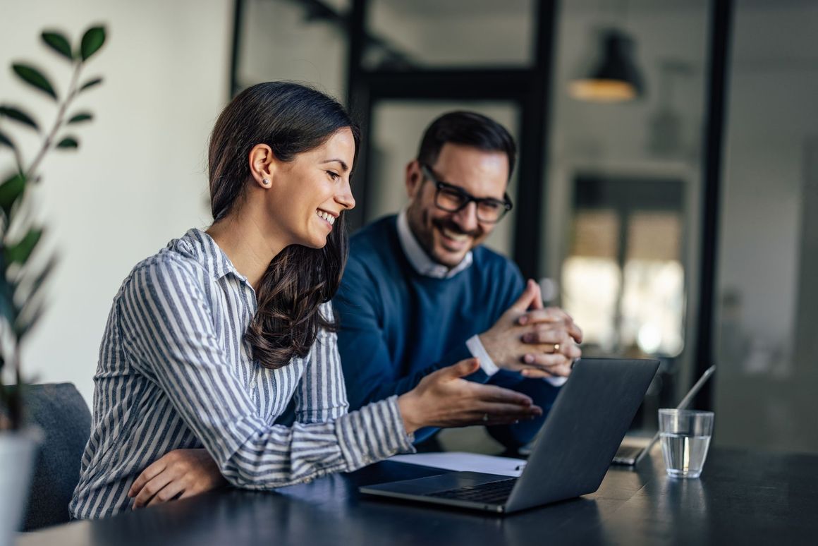 A man and a woman are sitting at a table looking at a laptop computer.