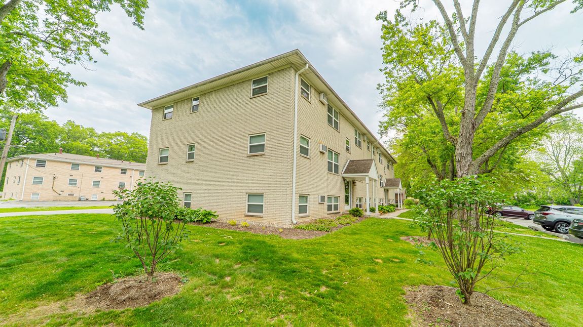 Three-story beige apartment building with multiple windows; green lawn and trees in the foreground.
