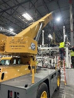 Technician on a ladder guiding industrial equipment into place with a mobile crane during a warehouse rigging installation.