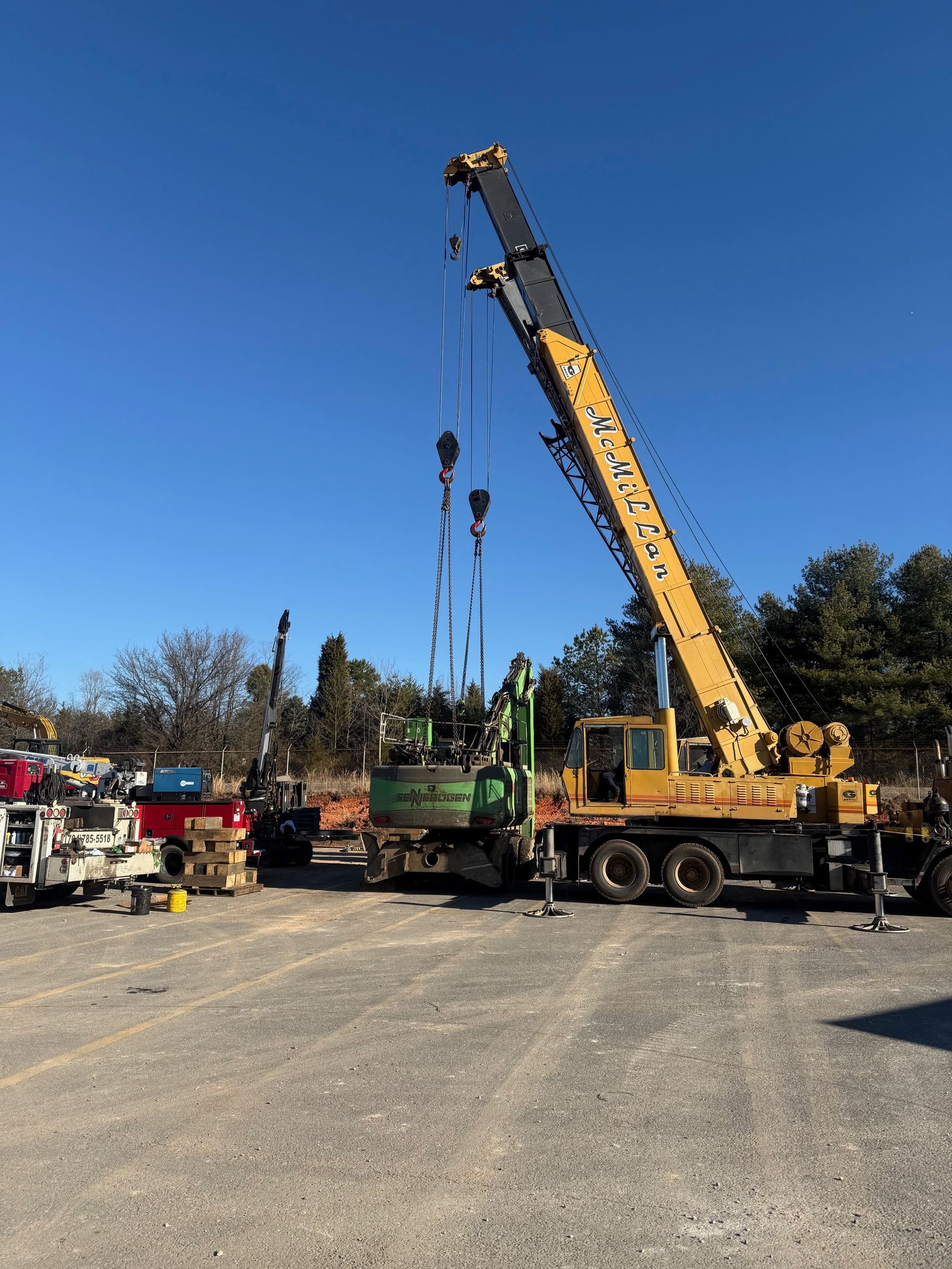 A large yellow crane parked on a gravel lot under a clear blue sky, with construction equipment nearby.