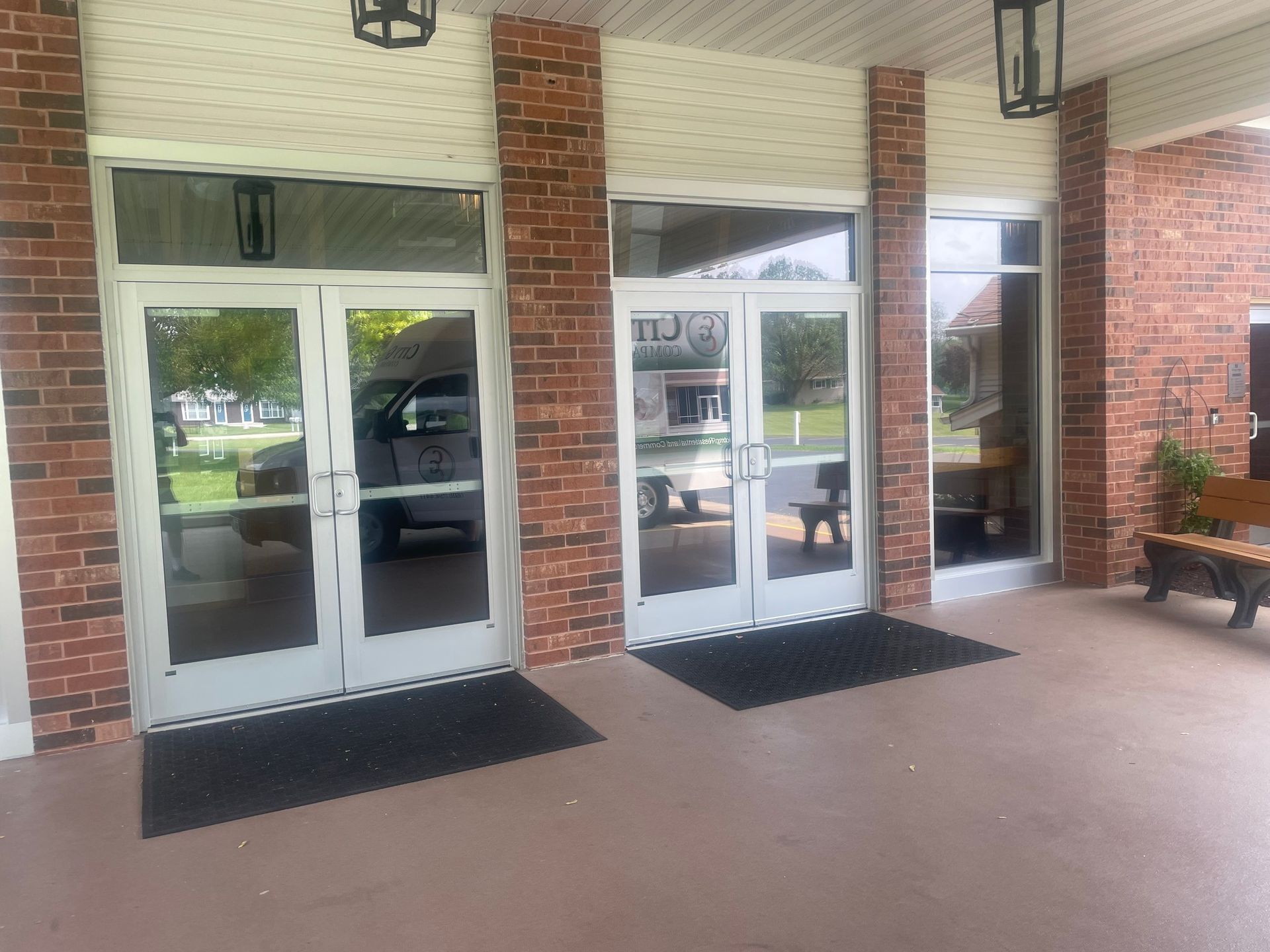 Exterior view of a building with three glass doors and brick columns, topped with hanging lanterns, and black doormats.