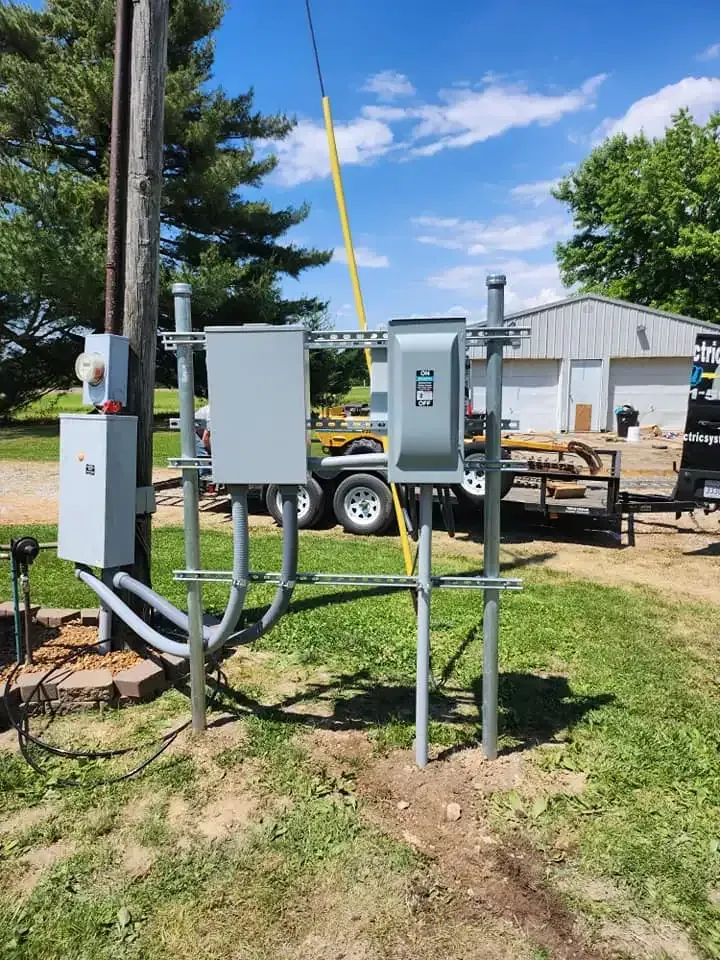 Electrical equipment mounted on metal poles, including grey boxes, conduits, and a meter, in a grassy, sunny outdoor area.