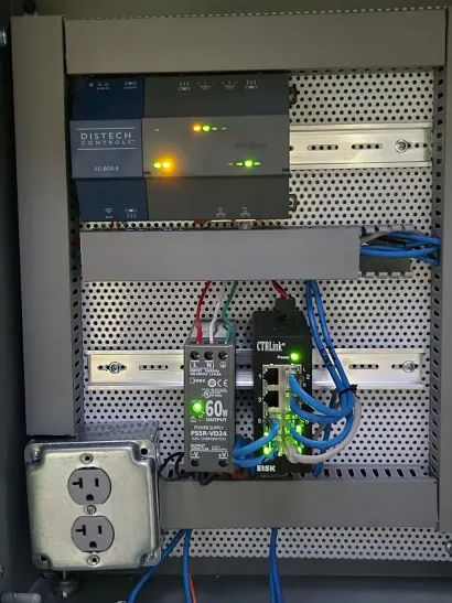 An industrial electrical enclosure containing a PLC, a power supply, a network switch, and a duplex power outlet.