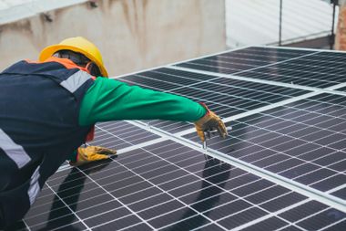 A worker in a yellow hard hat and green shirt installs solar panels on a rooftop.