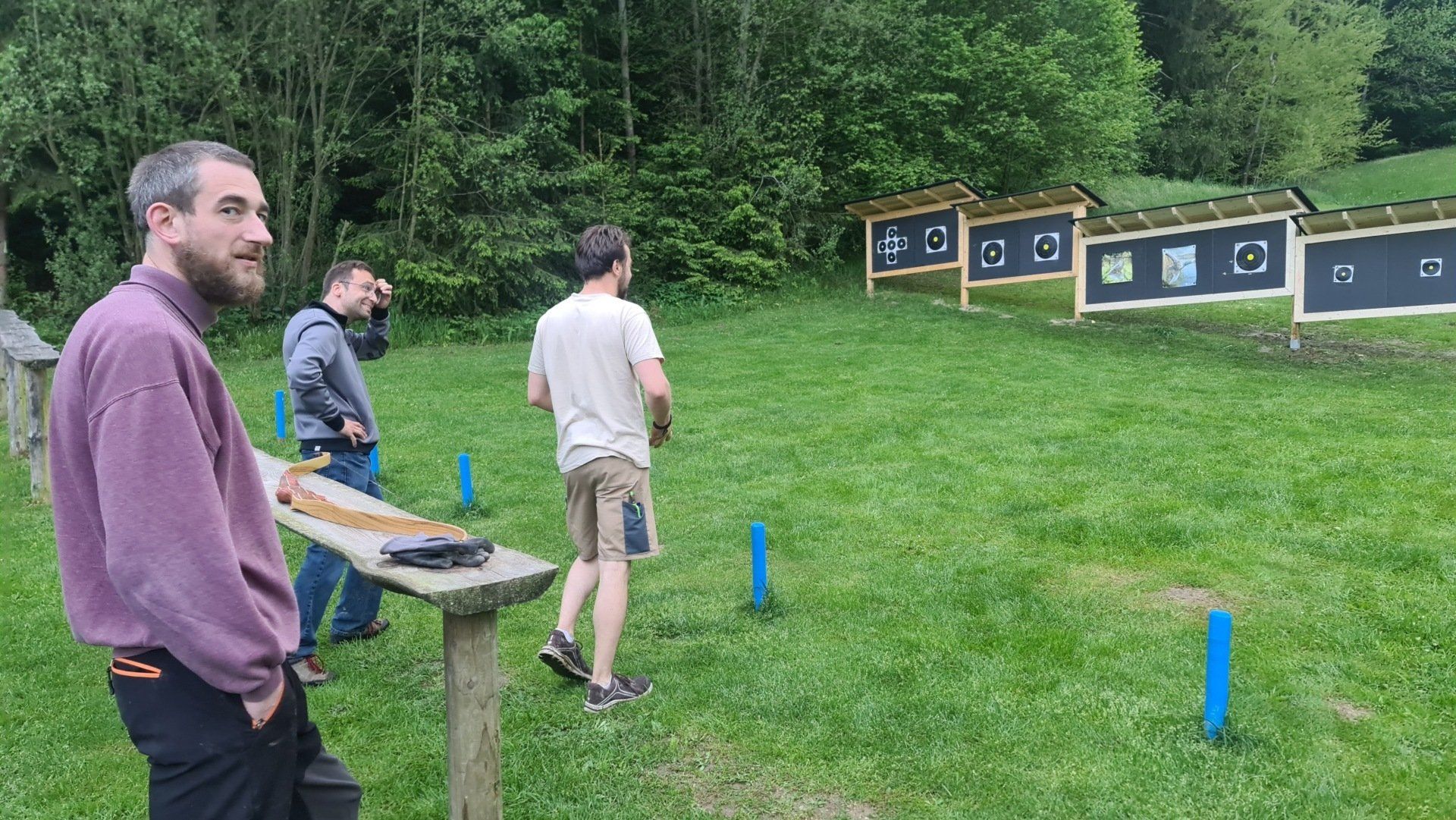 Drei Männer im Freien, bereiten sich auf das Zielschießen vor. Einer schaut zu, die anderen stehen bereit. Grünes Gras, Bäume im Hintergrund.