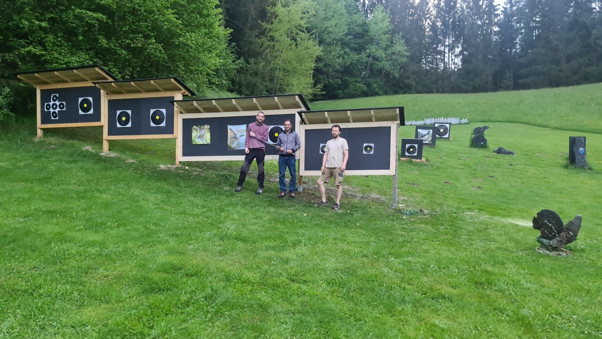 Drei Personen posieren vor Bogenschießscheiben auf einer grasbewachsenen Wiese in einem Waldgebiet.