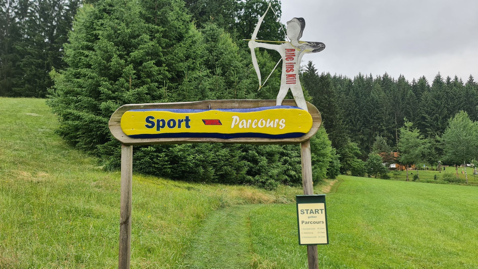 Schild mit der Aufschrift „Sport Parcours“ mit einem ausgeschnittenen Bogenschützen auf einem Holzrahmen in einer grasbewachsenen Wiese.