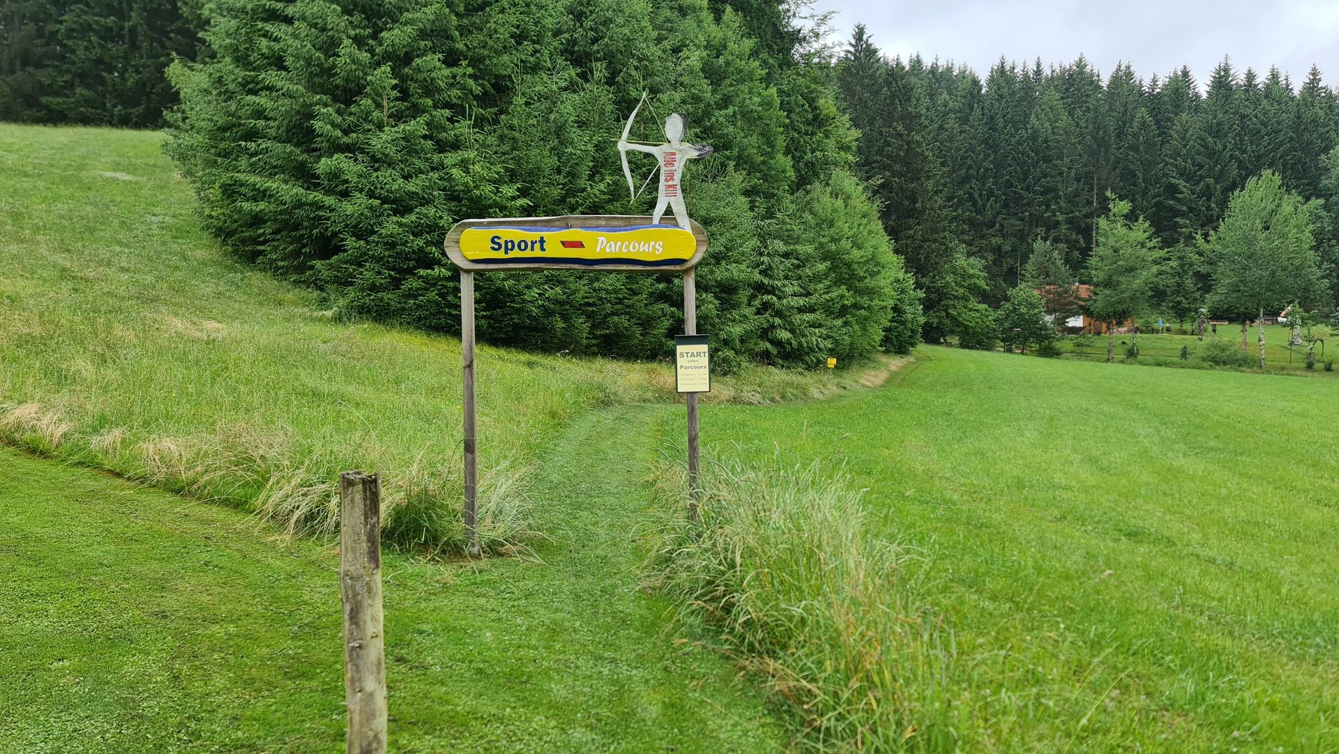 Holzschild mit Bogenschützenfigur, Pfad durch eine grasbewachsene Wiese. Wald im Hintergrund.