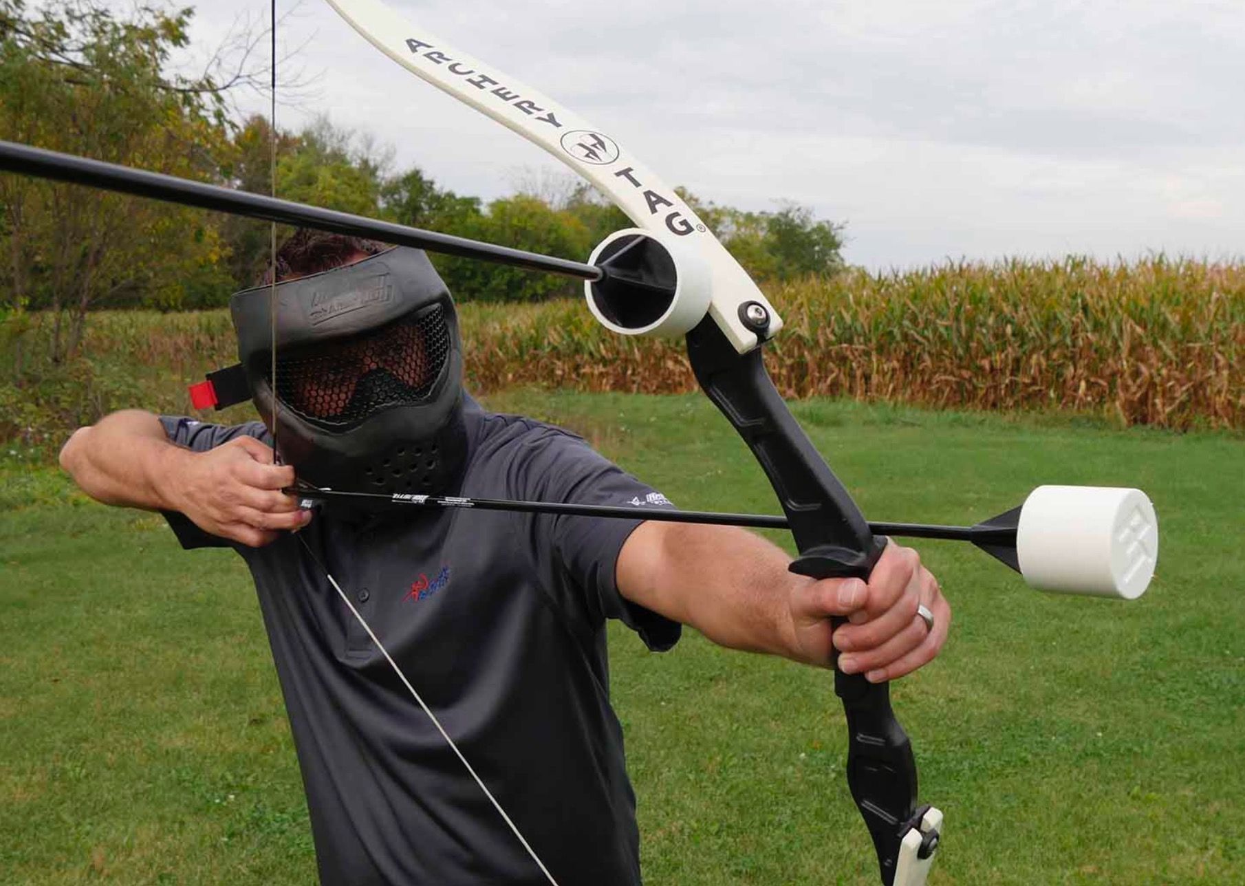 Ein Mann mit Paintballmaske zielt im Freien in der Nähe eines Feldes mit einem Spezialbogen, der über eine weiße, zylindrische Pfeilauflage verfügt.