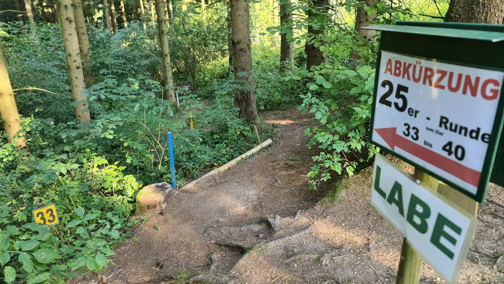 Ein Schild weist auf eine Abkürzung hin, die in den Wald führt. Es zeigt die runden Hausnummern 25, 33–40 an. Darunter befindet sich ein Schild mit der Aufschrift „LABE“.
